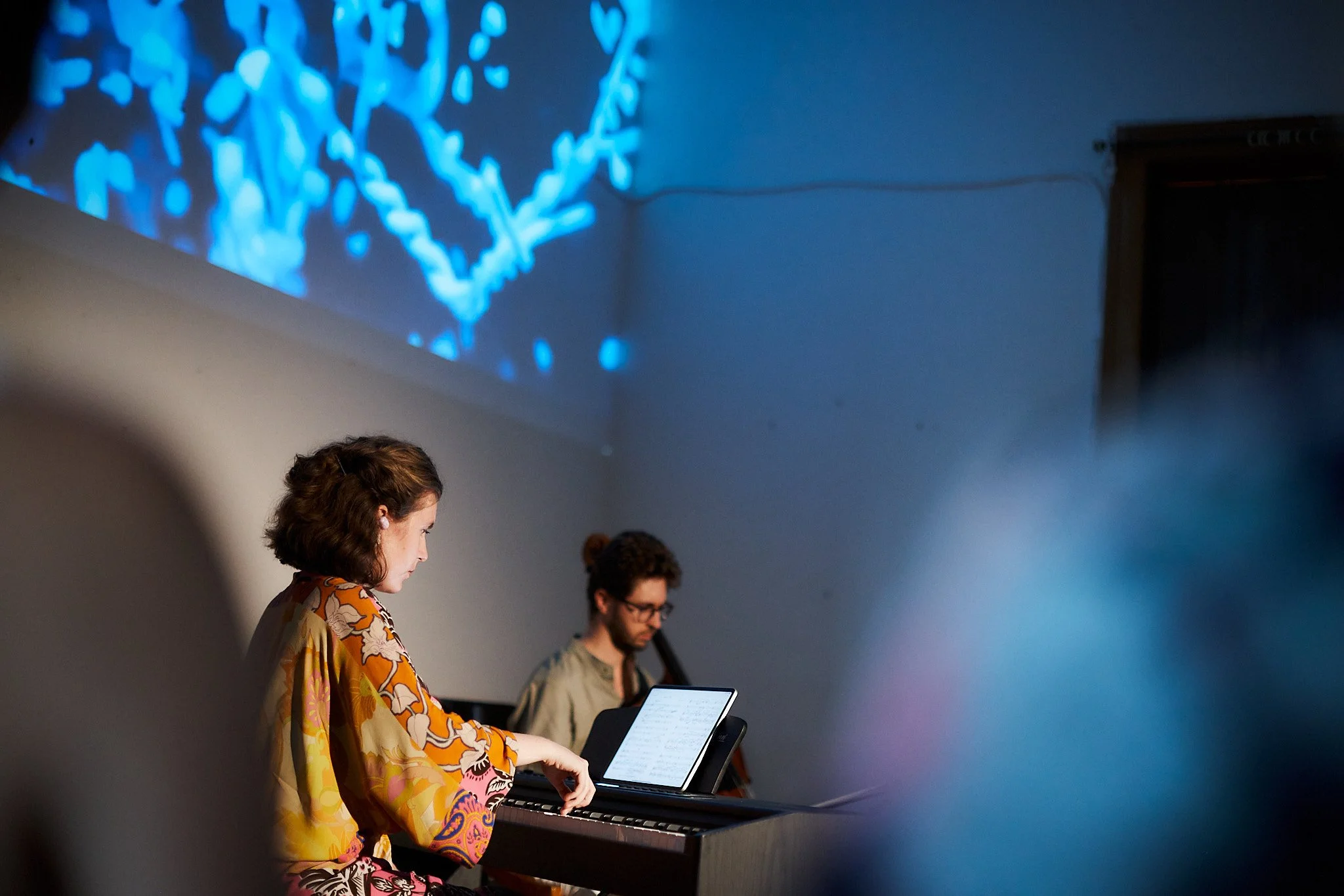 Two musicians playing the piano, with a woman on the left and a man on the right. The woman is wearing a colorful patterned dress, and the man has glasses and curly hair. A music sheet is visible on the piano.