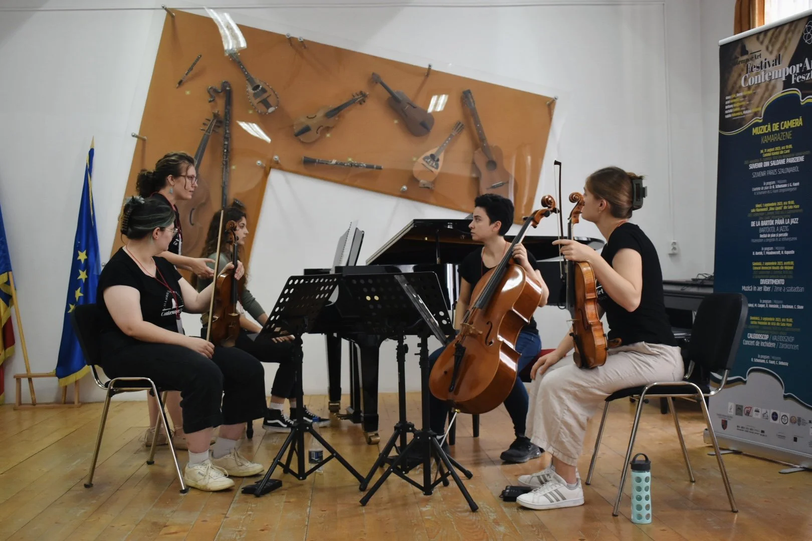 A group of five musicians rehearsing indoors, playing string instruments including violins, a cello, and a piano, with music stands in front of them. The room has a wooden floor and flags, with a large display of guitars mounted on the wall behind them.