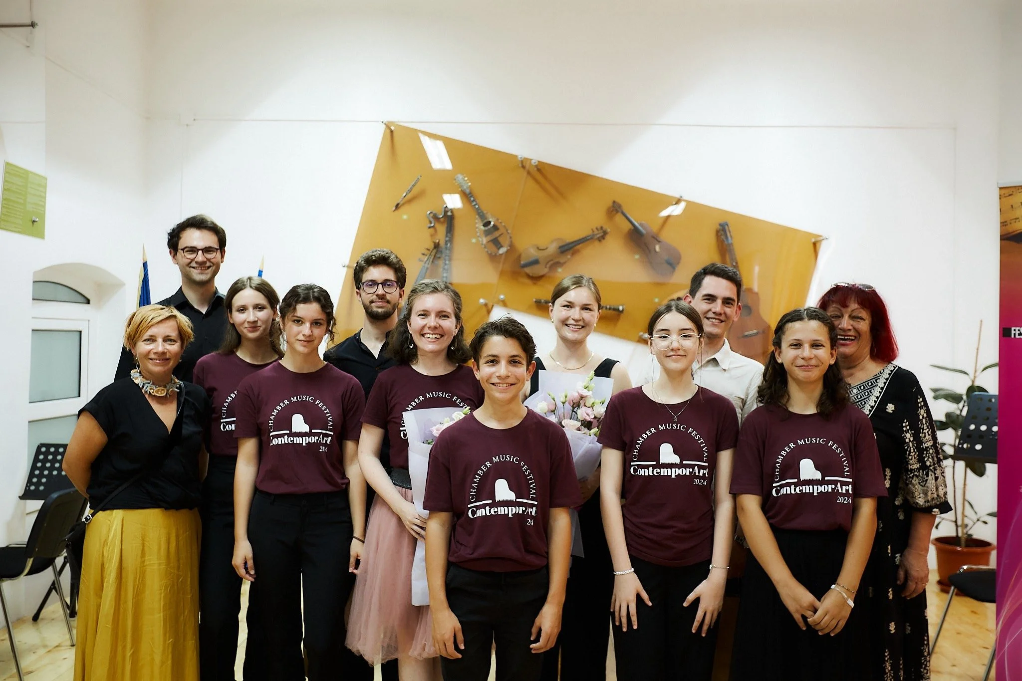 Group of young musicians and their teachers posing for a photo in a music practice room, some wearing maroon T-shirts with 'Contemporary Art 2024' printed on them, with musical instruments on the wall behind them.