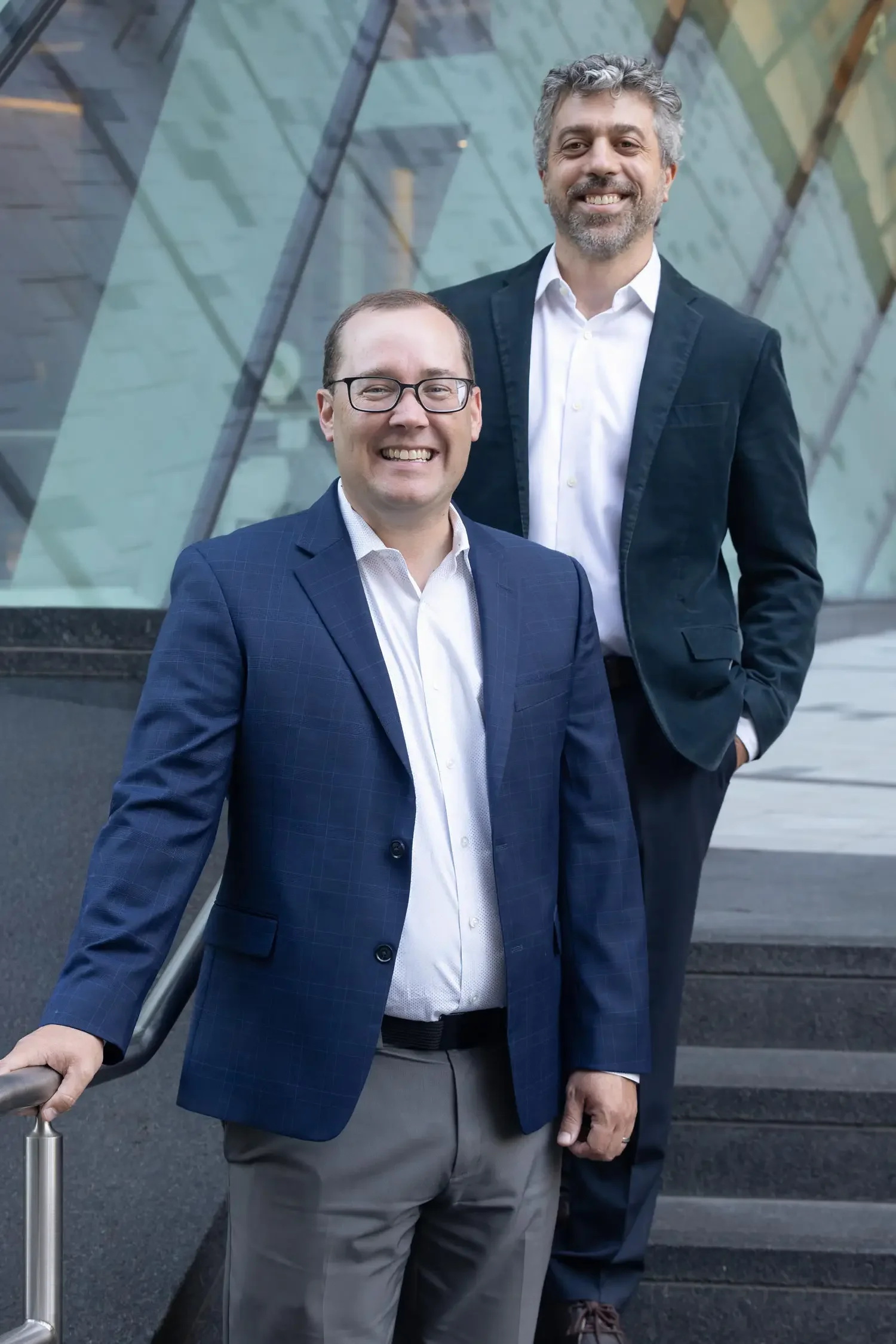 Steve Abreu and Peter Karol of Abreu Karol Trademark Law wearing business attire standing on steps in Boston Massachusetts outside a modern building with glass facade, smiling for the photo.