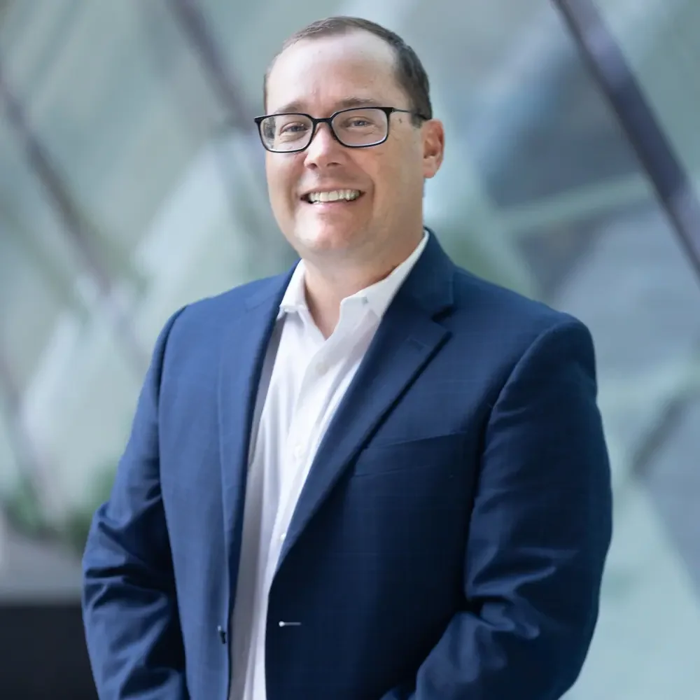 Steve Abreu of Abreu Karol Trademark Law in a navy blue suit and white shirt, wearing glasses, smiling and standing in front of a modern glass building.