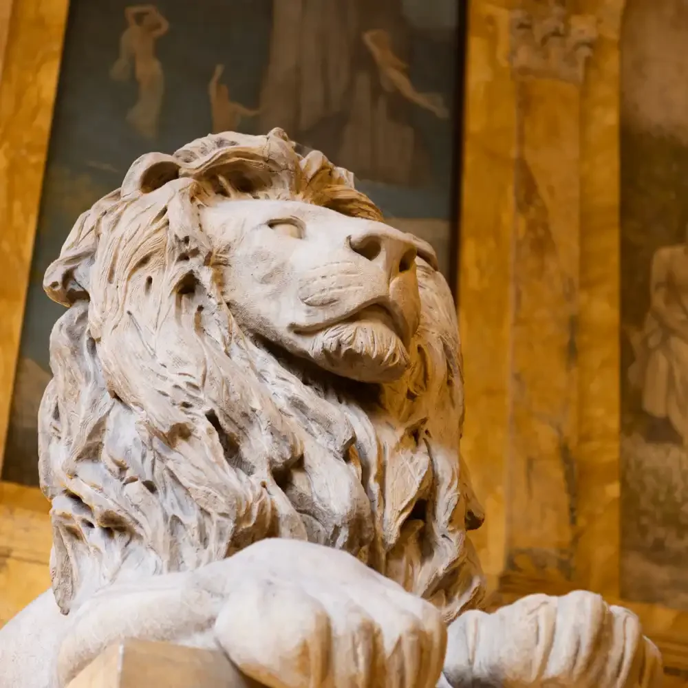 A detailed marble sculpture of a lion's head with a lush mane, displayed in the Boston Public Library with wooden paneling and framed artwork in the background.