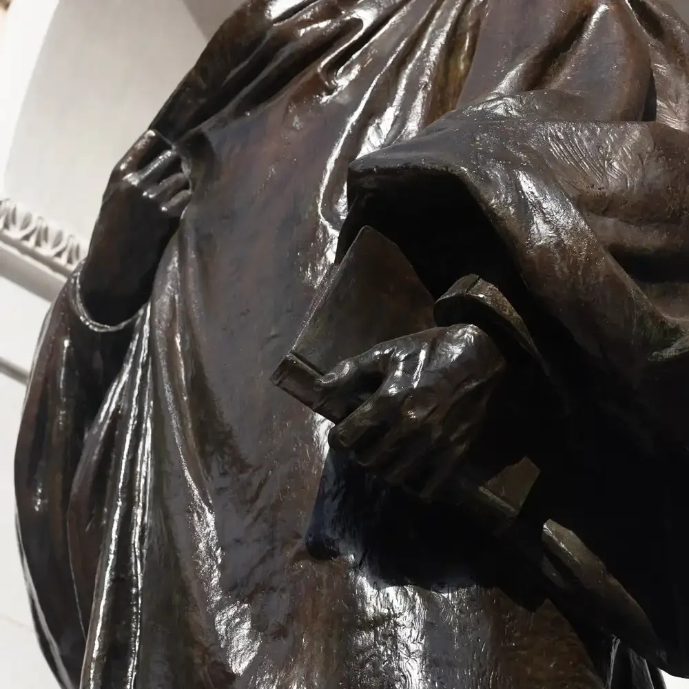 Close-up of a bronze sculpture showing a person's hand holding a book, with detailed folds in their clothing outside the Boston Public Gardens.