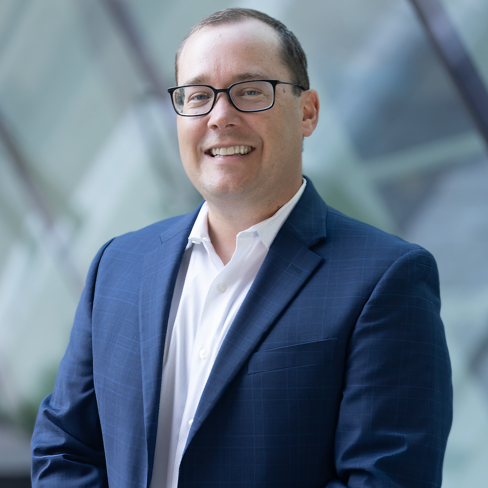 Steve Abreu of Abreu Karol Trademark Law in a blue suit and white shirt smiling, wearing glasses, standing in front of an abstract glass building background.