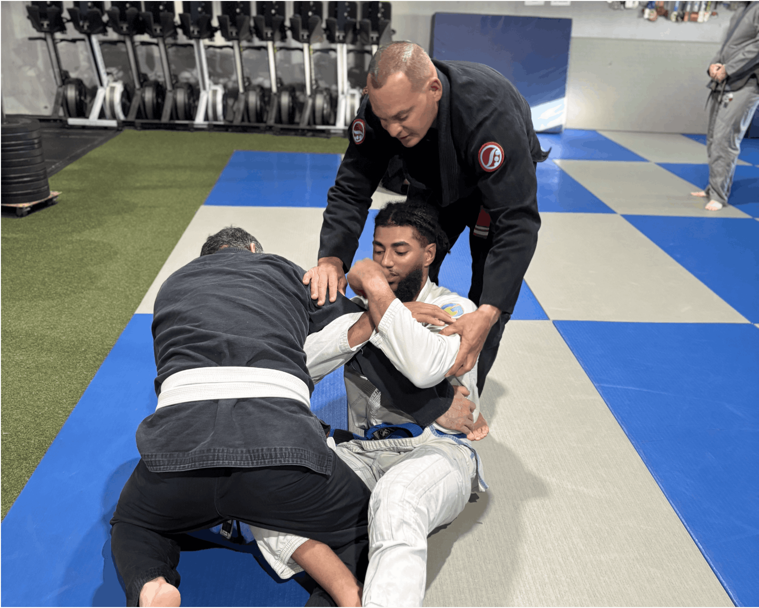 Two martial artists in Brazilian Jiu-Jitsu uniforms practicing a submission hold on a padded mat. One is on his back, while the other is leaning over him, and a third person is assisting or coaching.