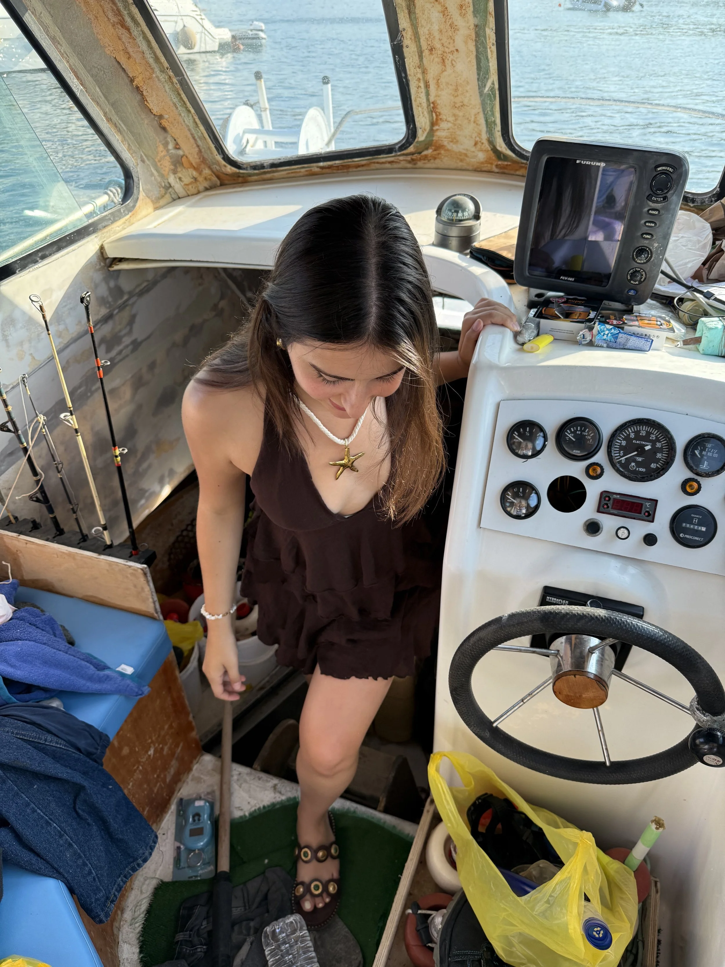 A woman standing inside a boat near the steering wheel, with a boat dashboard, fishing gear, and various small items around her. The boat is on water with lots of other boats visible outside through the windows.