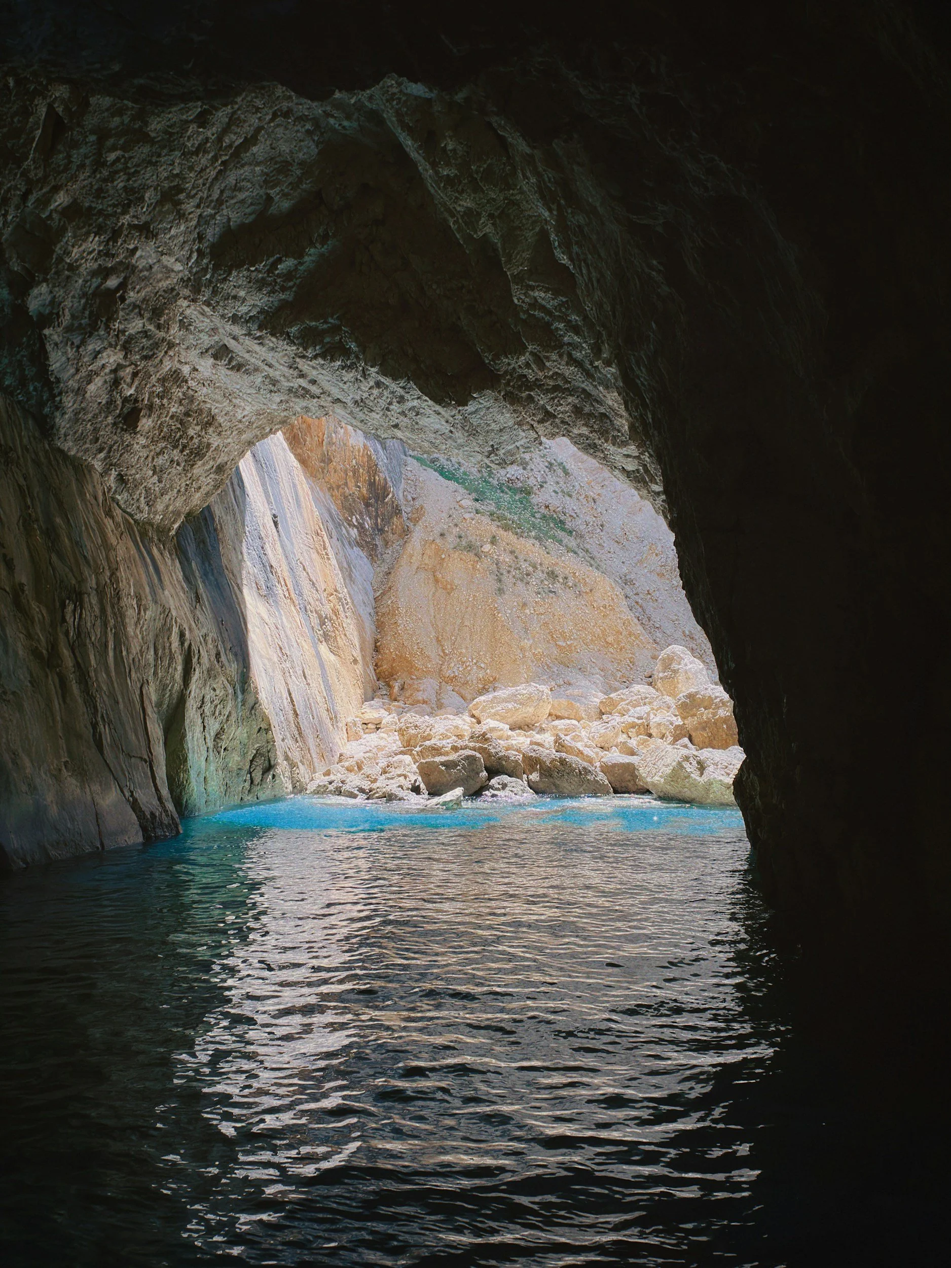 View from inside a dark cave looking out at a bright, sunny cove with turquoise waters, rocky cliffs, and a sandy shoreline.