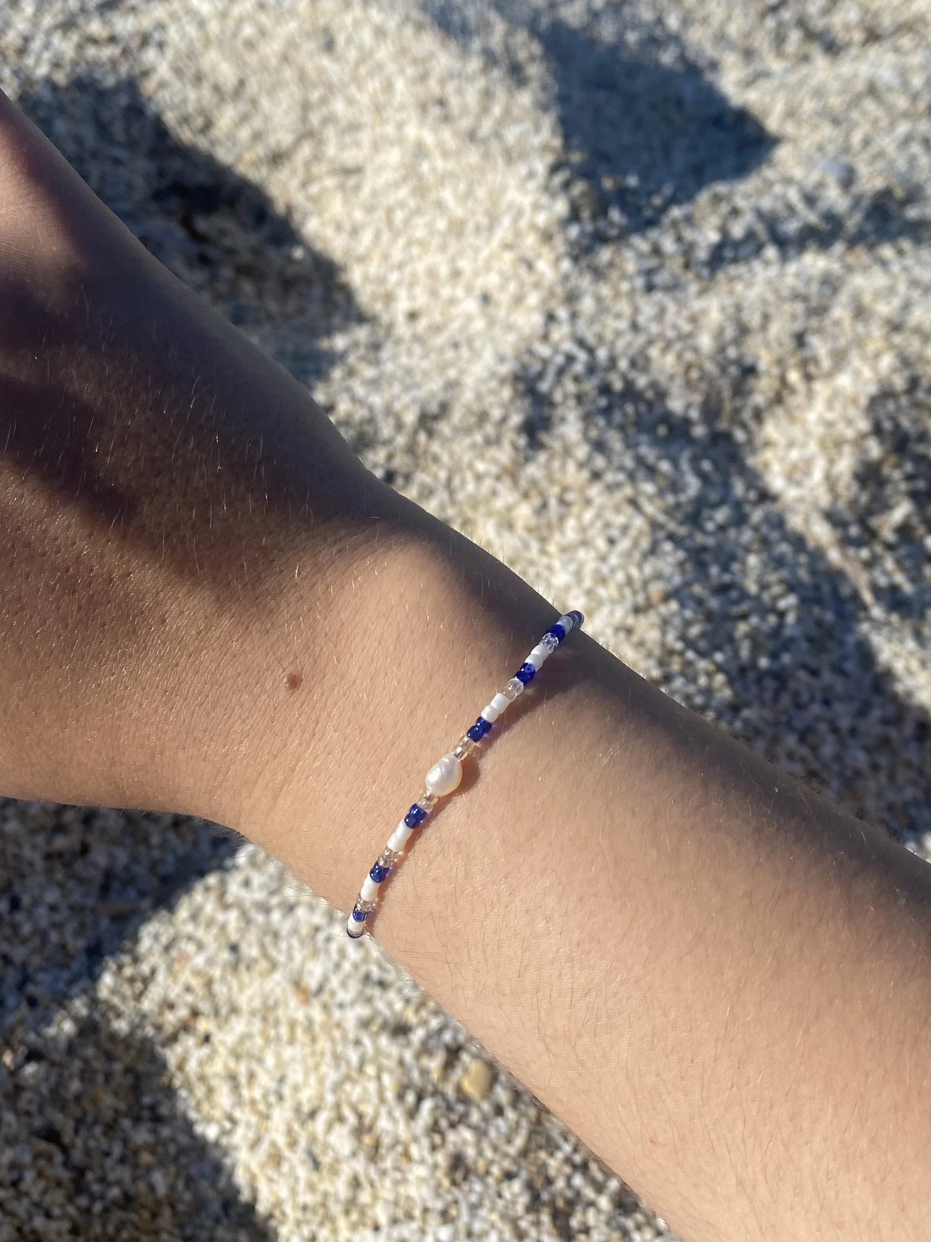 A person's arm wearing a beaded bracelet with blue, white, and pearl beads against a sandy ground with pebbles.