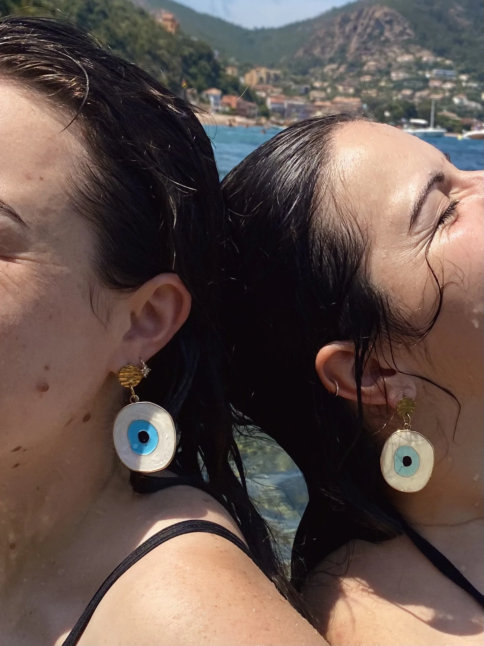 Close-up of two women relaxing on a beach, with their heads close together and their eyes closed, enjoying the sun. They are wearing earrings with evil eye pendants, and have dark hair. In the background, there are mountains, water, and houses.