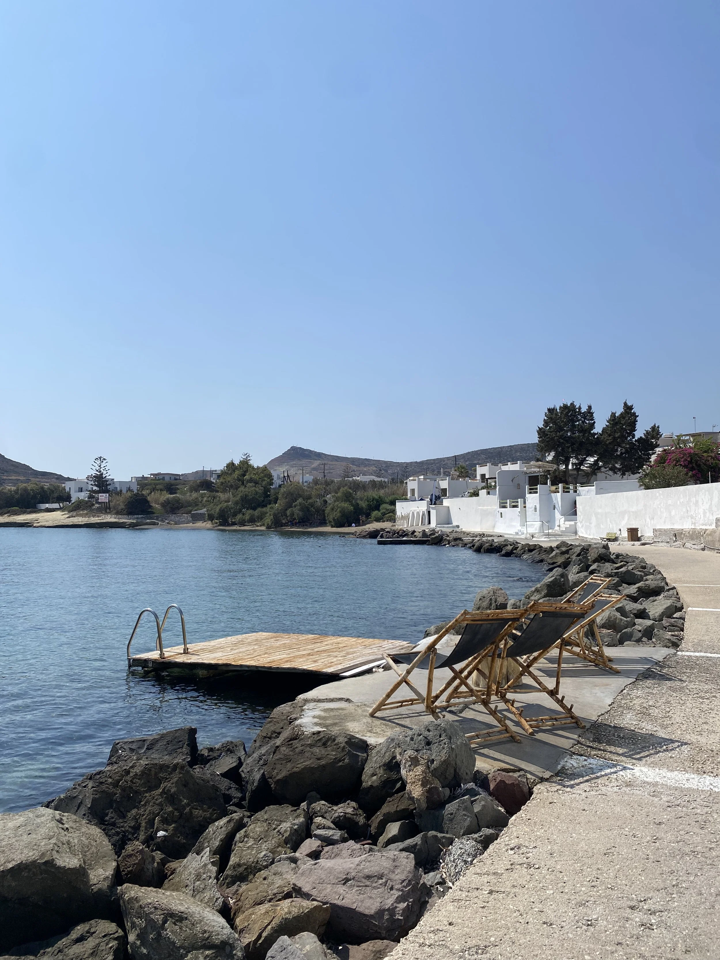 A lakeside scene with two lounge chairs on a concrete and rock shoreline, a wooden dock extending into the water, white buildings with flat roofs and trees in the background, and mountains under a clear blue sky.