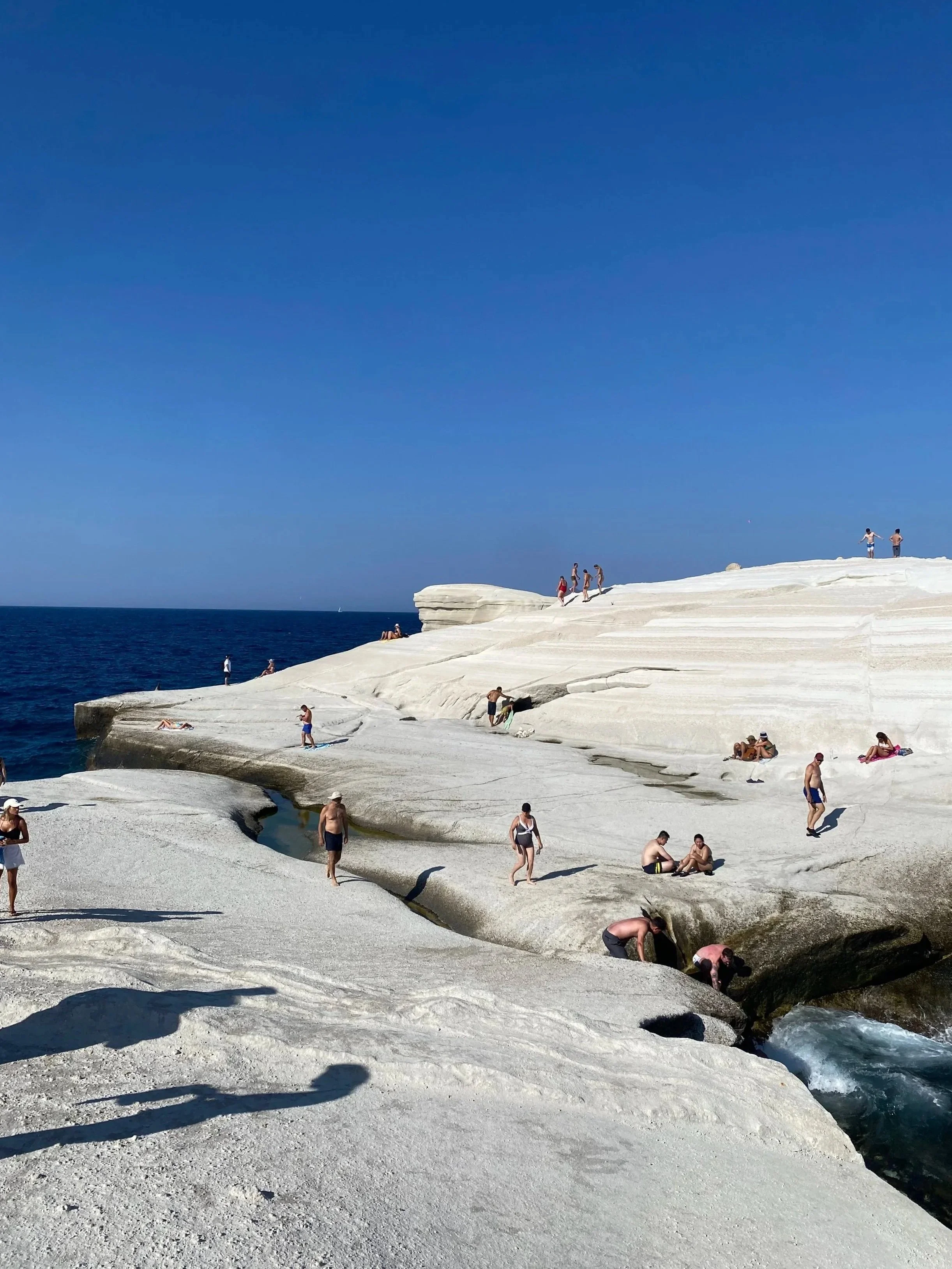 People relaxing and exploring on white rocky terrain by the sea under a clear blue sky.