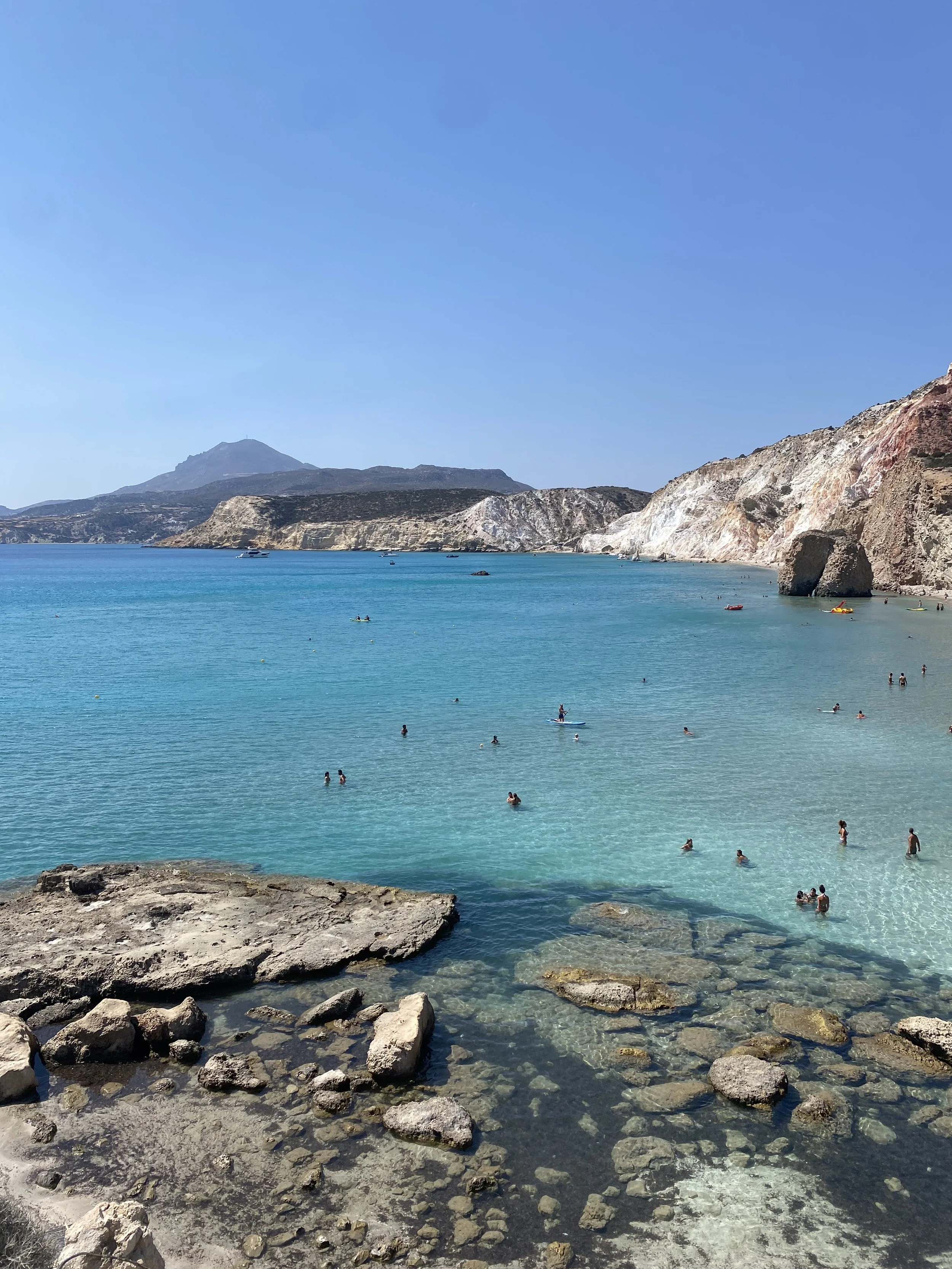 A scenic beach with clear turquoise water, people swimming and paddleboarding, rocky shoreline, and hills in the background under a bright blue sky.