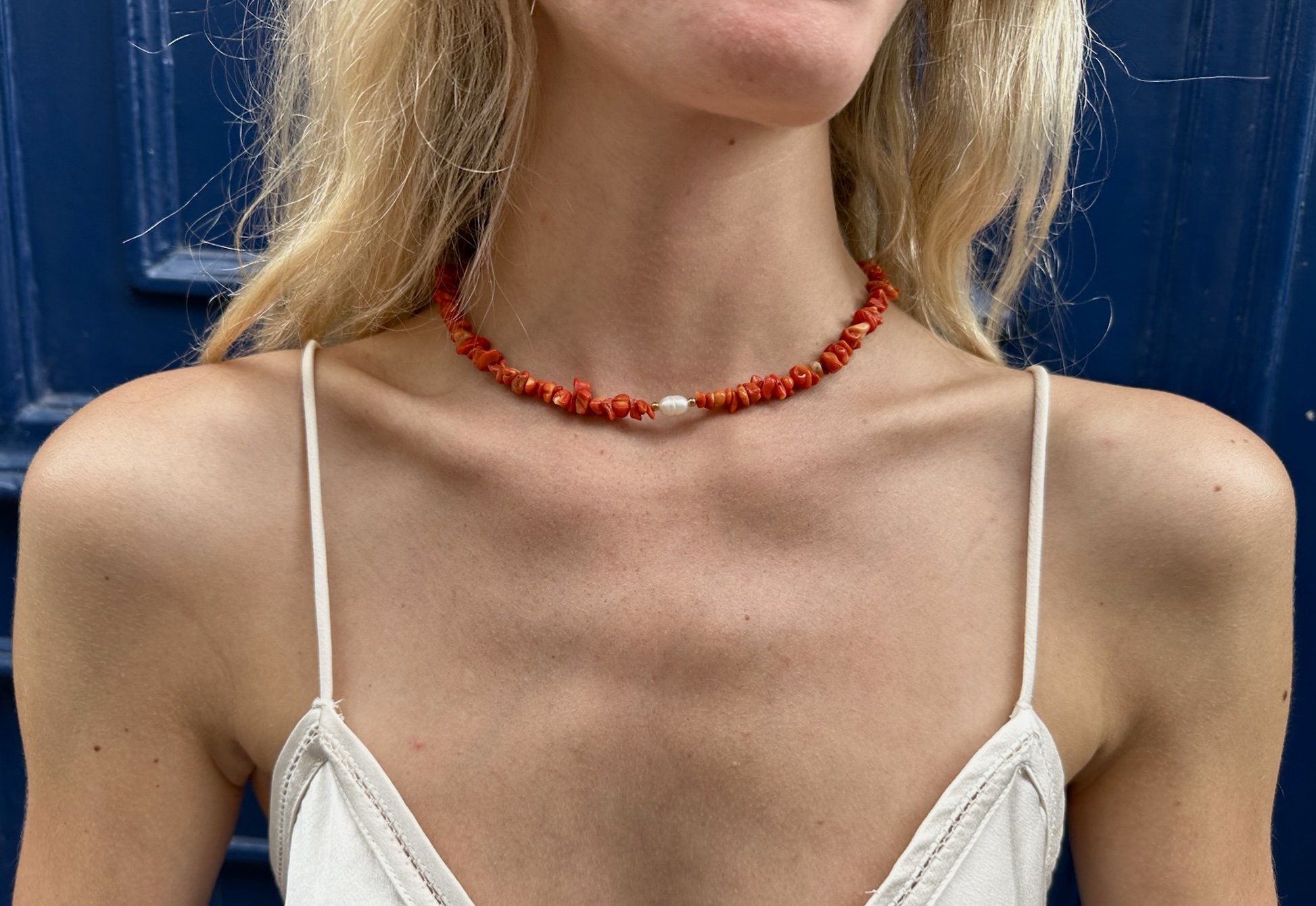 A woman wearing a red beaded necklace with a single white pearl in the center. She is dressed in a white spaghetti strap top and is standing in front of a blue wooden background.