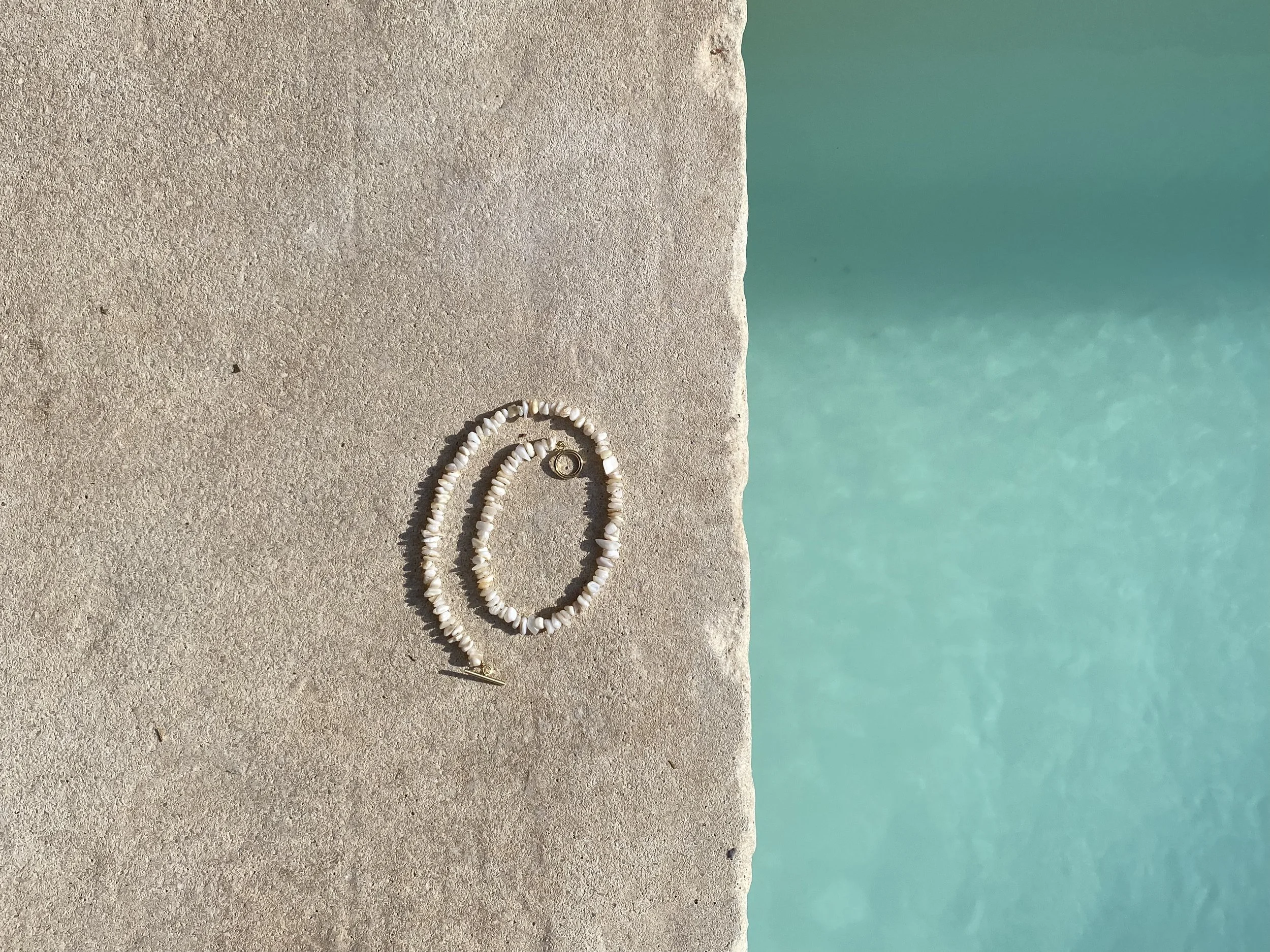 A pair of white beaded earrings with a gold clasp and a small gold charm, placed on a concrete surface near a turquoise-colored pool.