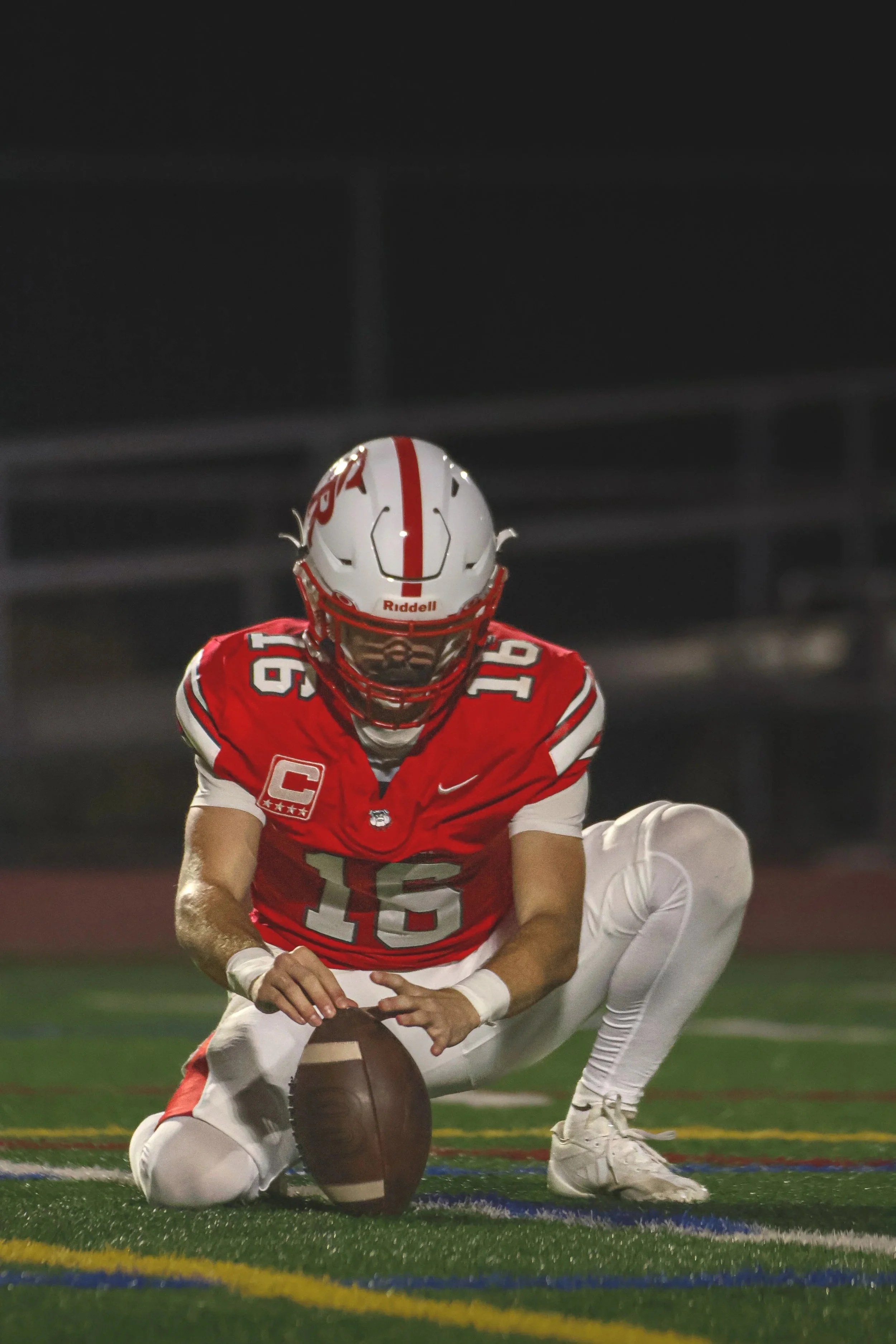 A football player in red and white uniform kneeling on the field, holding a football, moments before a snap or kick.