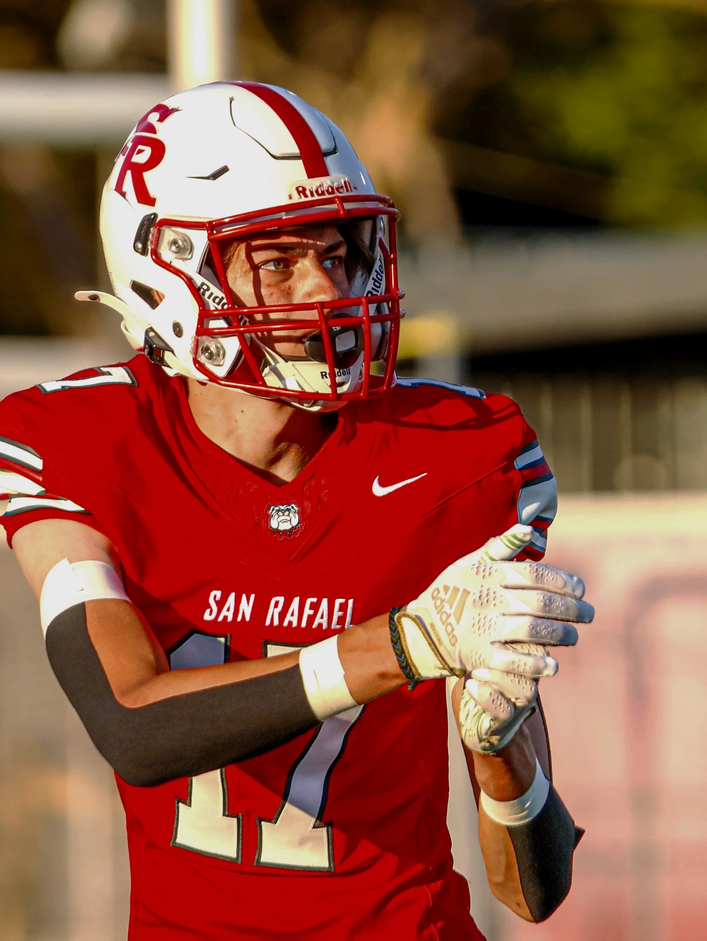 A football player wearing a red uniform with 'San Rafael' on the front, a white helmet with red detailing, and gloves, appears to be on the field during a game.