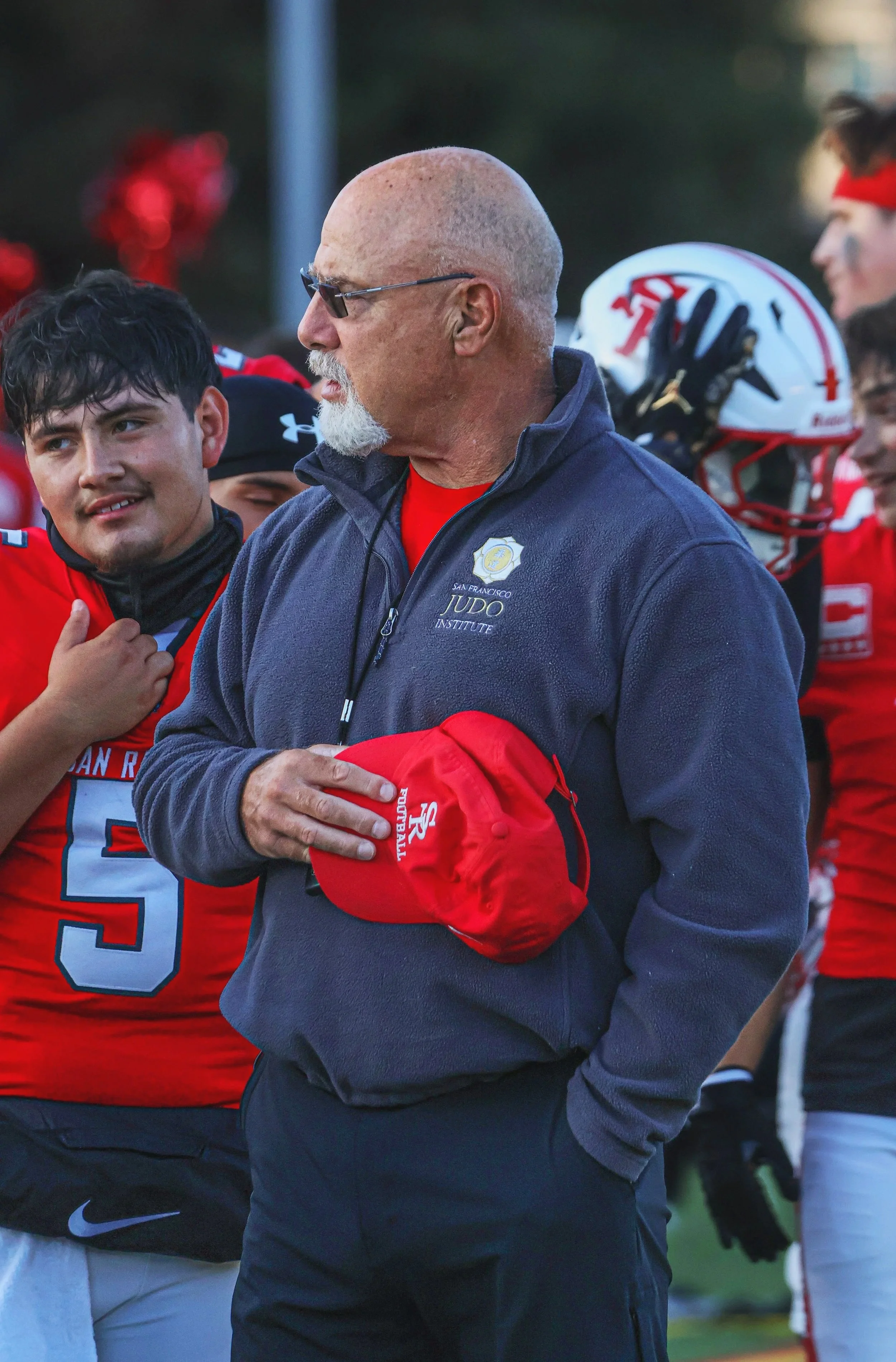 A man in sunglasses, a navy jacket with a San Francisco Judo badge, holding a red San Francisco Football cap, stands among football players in red uniforms during a game or practice.