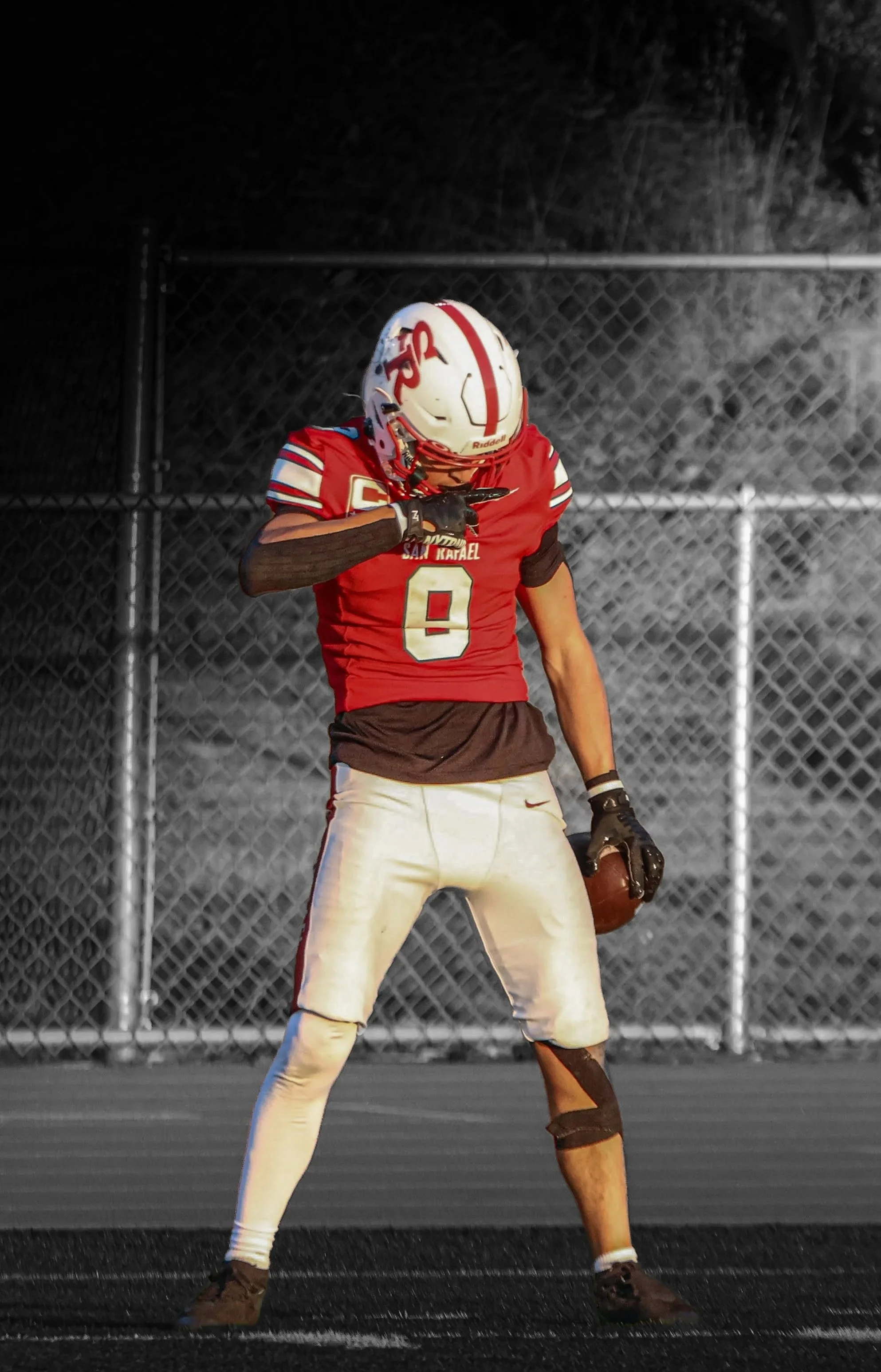 A football player in red jersey, white pants, and helmet standing on a football field at night, holding a football in one hand and touching his face with the other.