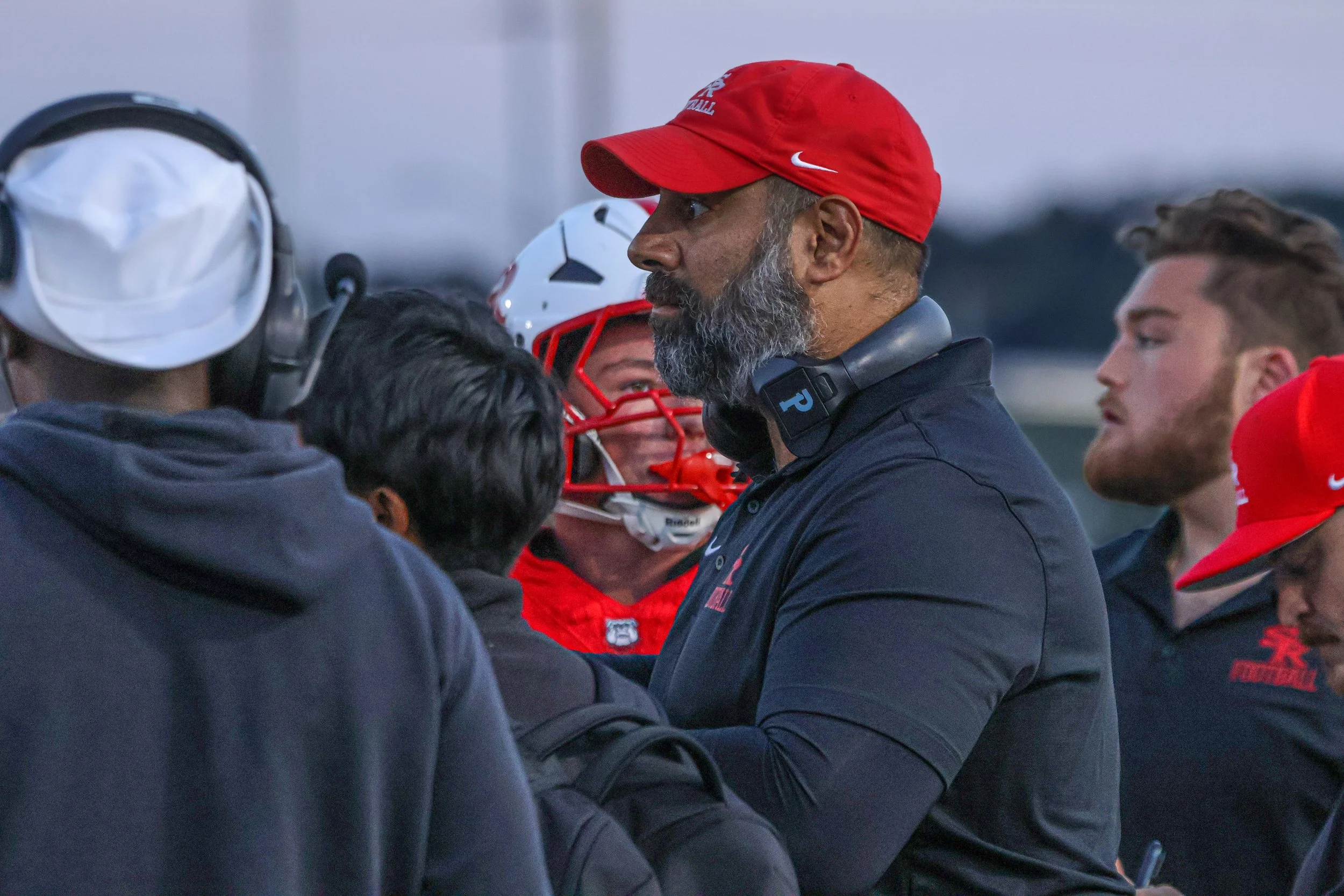 A coach in a black shirt and red cap speaking to a group of football players, some wearing helmets and red hats.