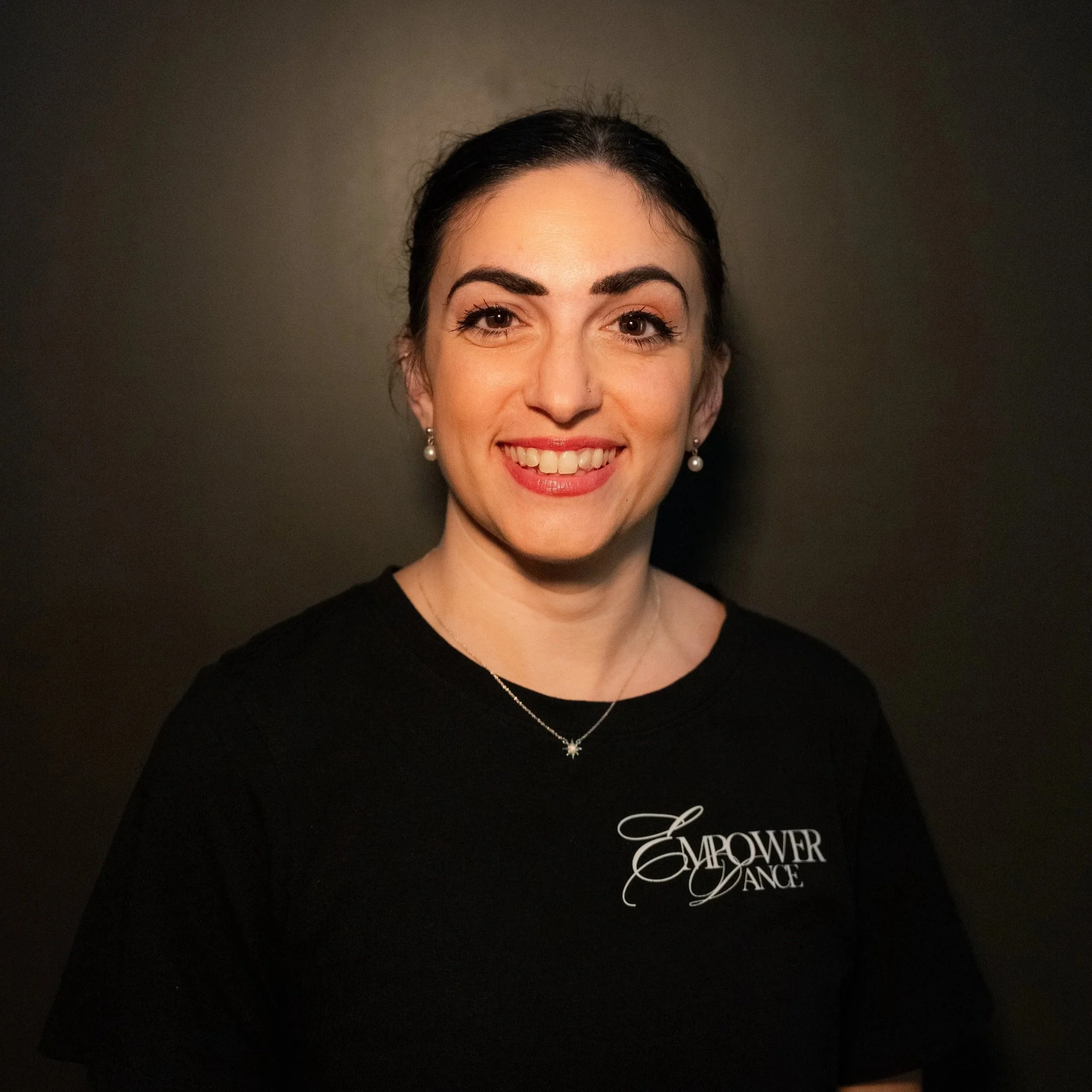A young woman with curly black hair smiling, wearing a white t-shirt, against a beige background.