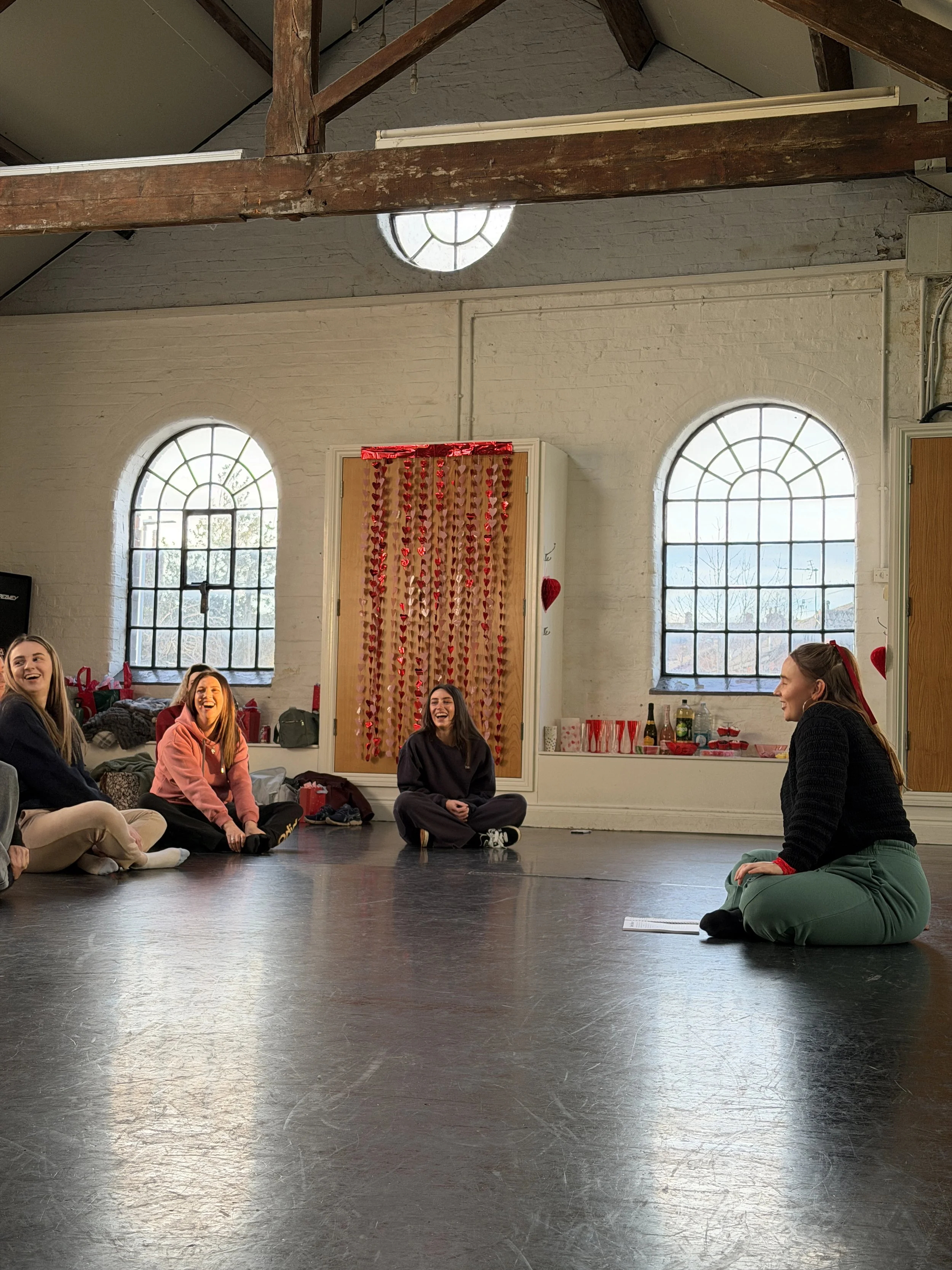Five women sitting on the floor in a circle inside a room with large arched windows, decorated with Valentine's Day hearts and red decorations.