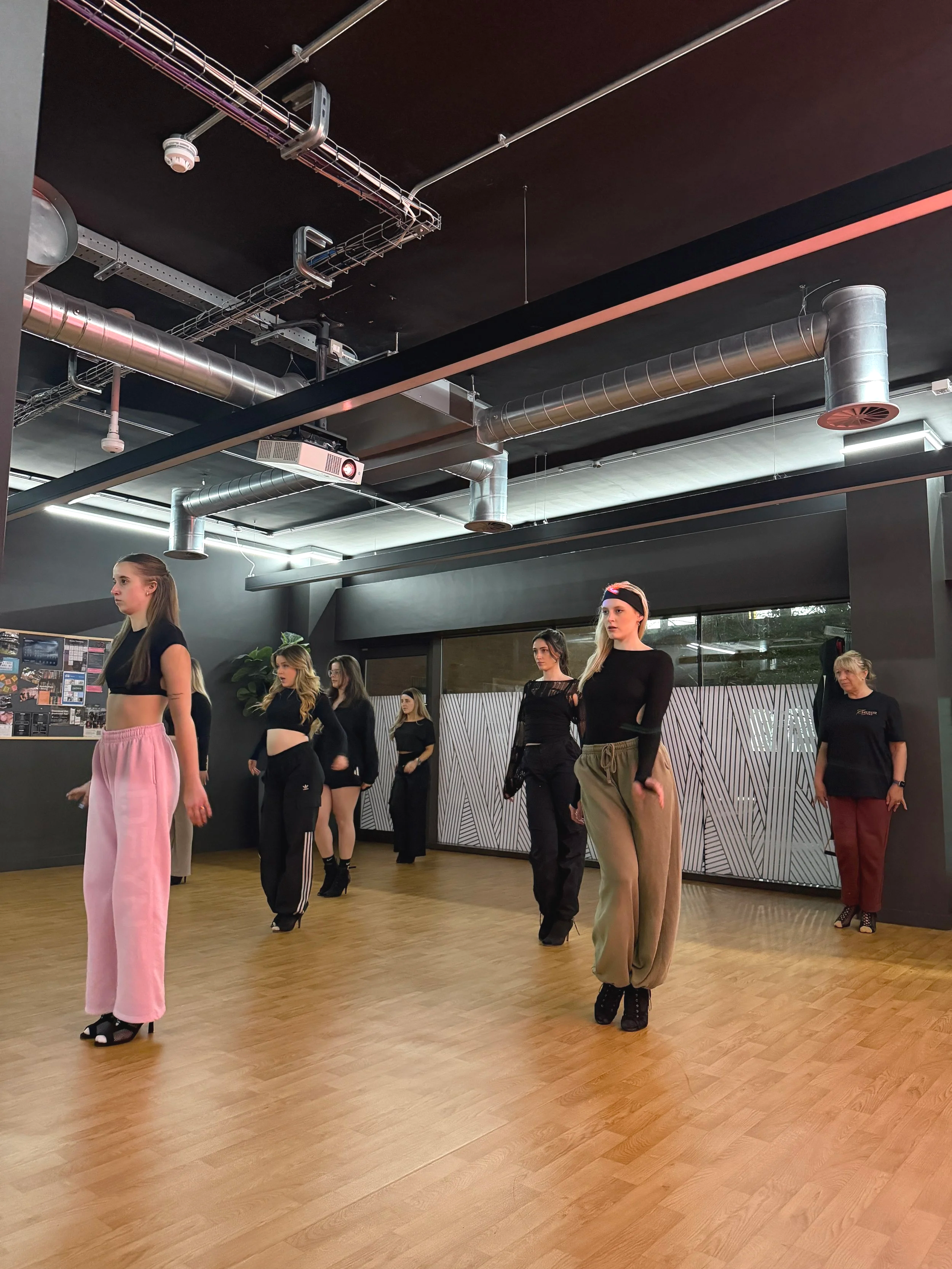 Group of women in stylish dance or exercise clothing practicing in a dance studio with wooden floors and modern industrial ceiling.
