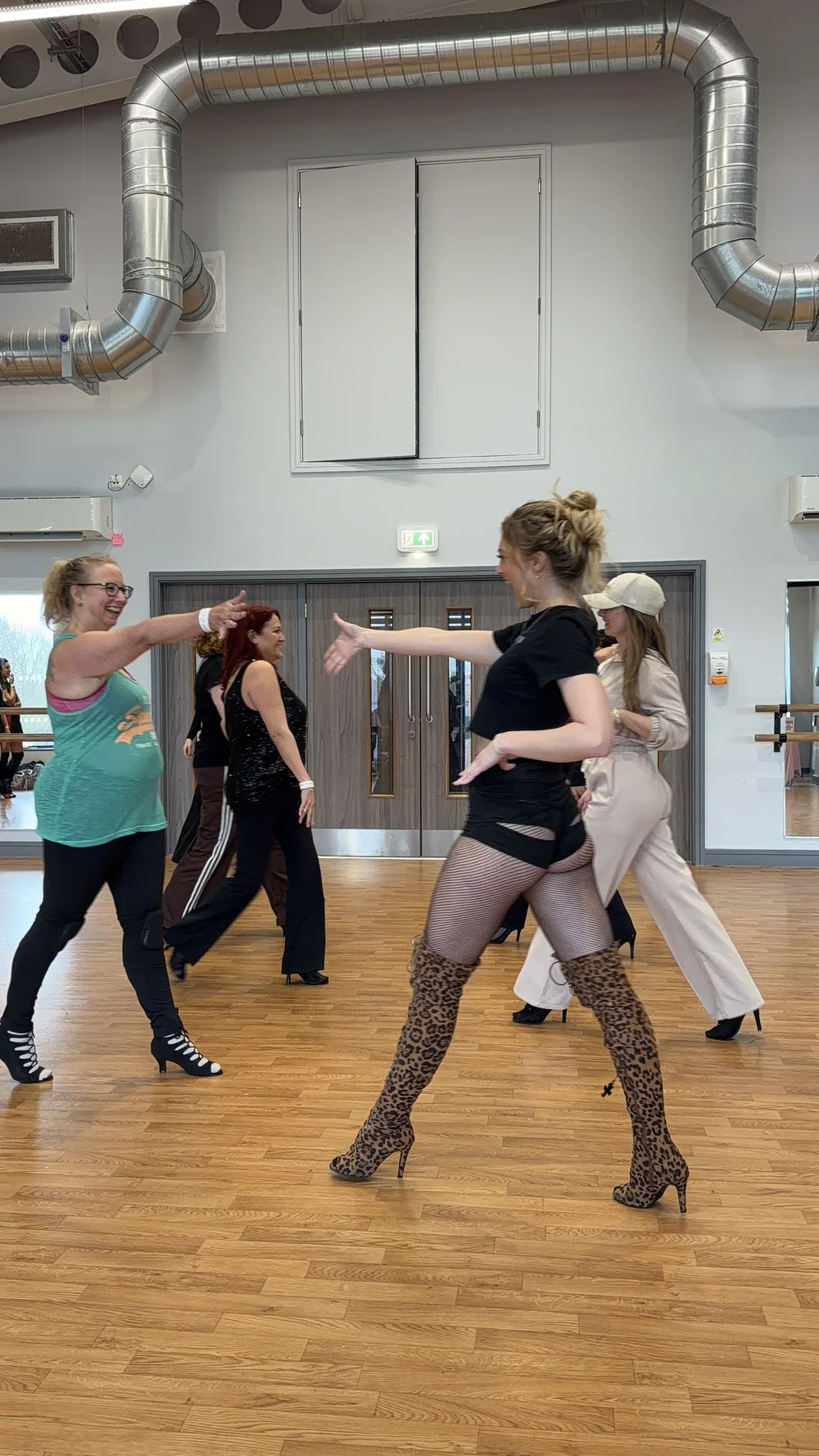 Group of women dancing in a studio with wooden floors, with some high-heeled boots and various outfits, including fishnet stockings and animal print thigh-high boots.