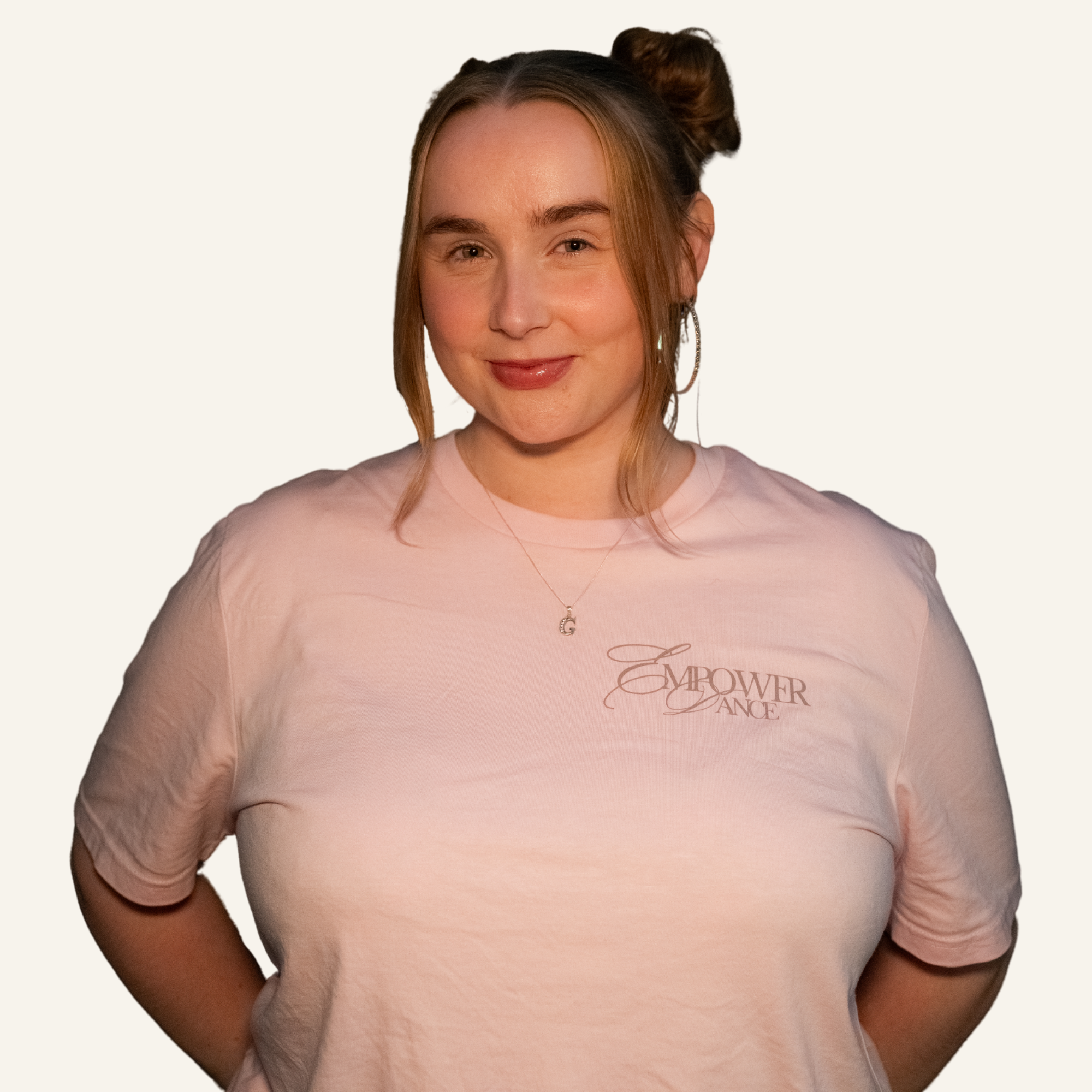 A young woman with light skin and brown hair styled in buns, wearing a light pink T-shirt with 'Empower Dance' written on it, a necklace, and hoop earrings, smiling at the camera against a plain dark background.