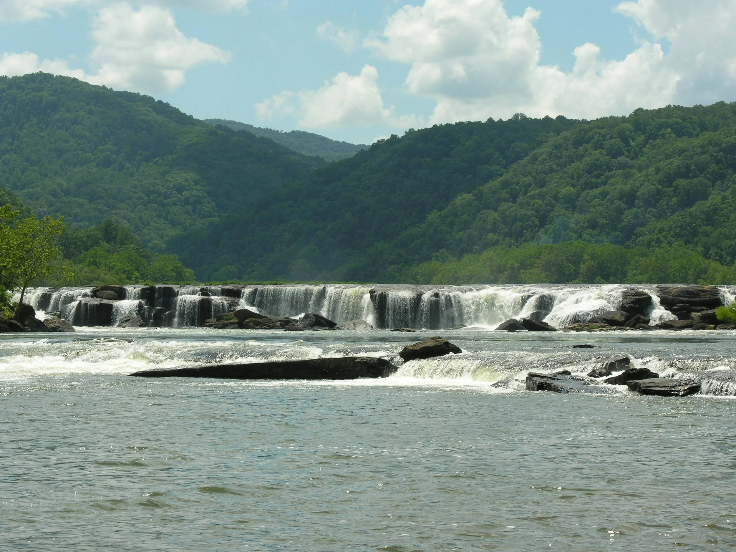 A scenic view of a river with a rock waterfall, surrounded by green mountains under a partly cloudy sky.