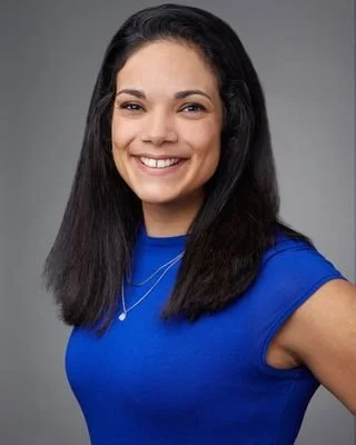 A young woman with dark hair wearing a blue shirt and smiling at the camera against a gray background.