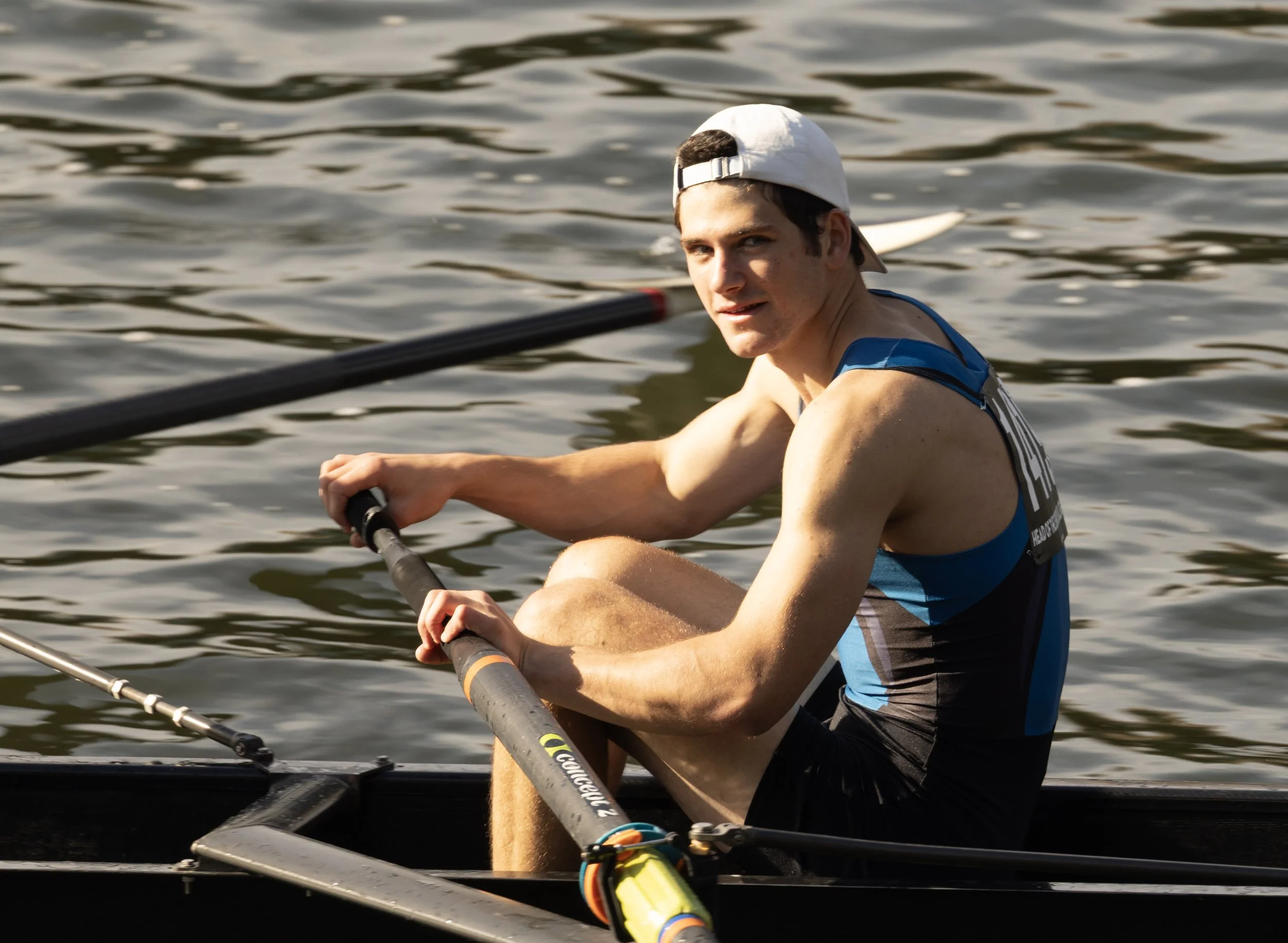 A young male rower wearing a white cap and sports gear, sitting in a rowing boat on the water, holding an oar and looking at the camera.