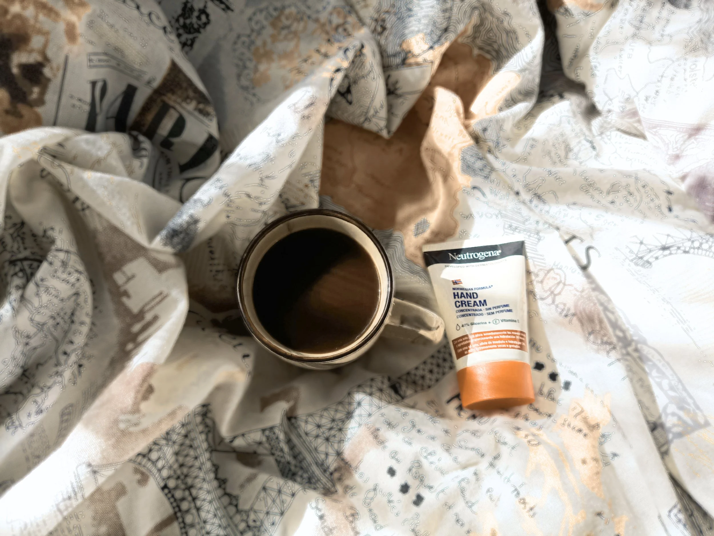 A cup of black coffee, a tube of Neutrogena hand cream, and a crumpled sheet of paper with a map and writing are placed on a surface.