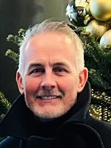 A smiling man with gray hair and a beard standing in front of a decorated Christmas tree with gold and silver ornaments.