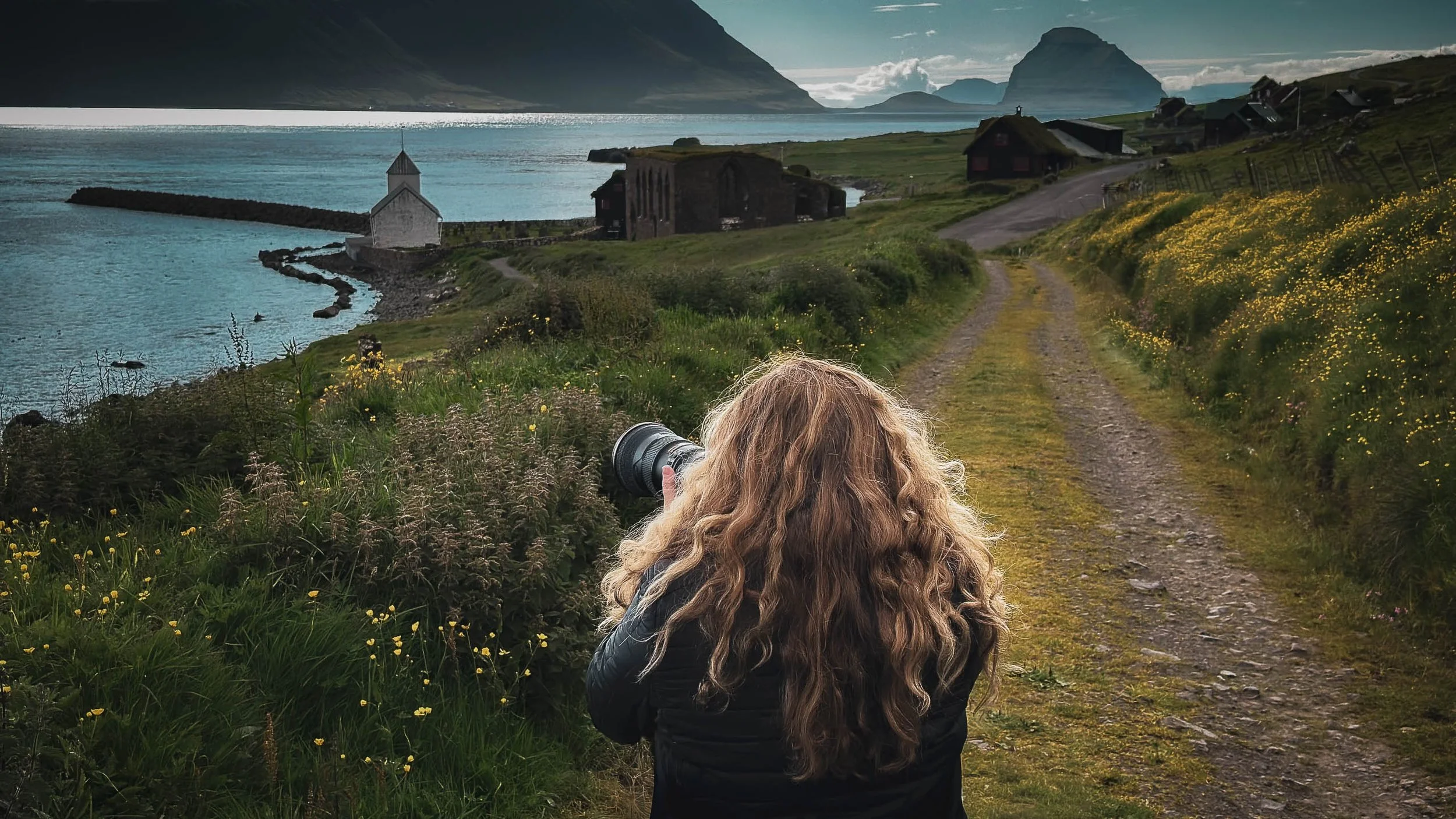 A woman with long curly hair taking a photograph with a camera on a grassy trail overlooking the Faroe Islands, with mountains and a cloudy sky in the background.