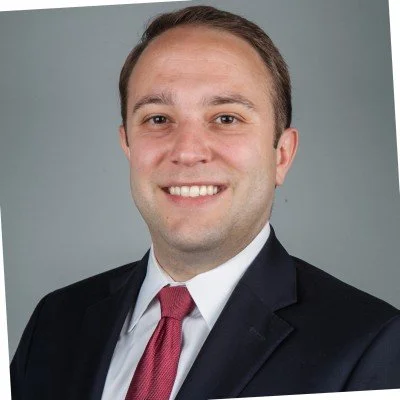 Portrait of a smiling man in a dark suit, white shirt, and red tie against a gray background.