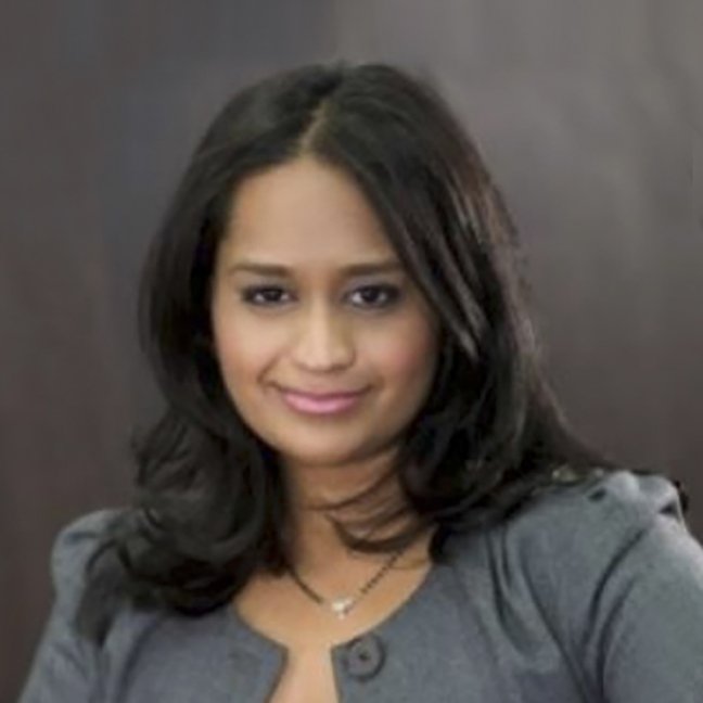 A woman with shoulder-length dark hair, smiling, wearing a gray blazer and layered necklaces, against a dark background.