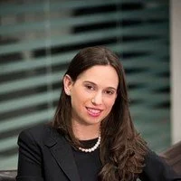 Professional woman with long dark hair, wearing a black blazer and pearl necklace, smiling in an office setting with blinds in the background.