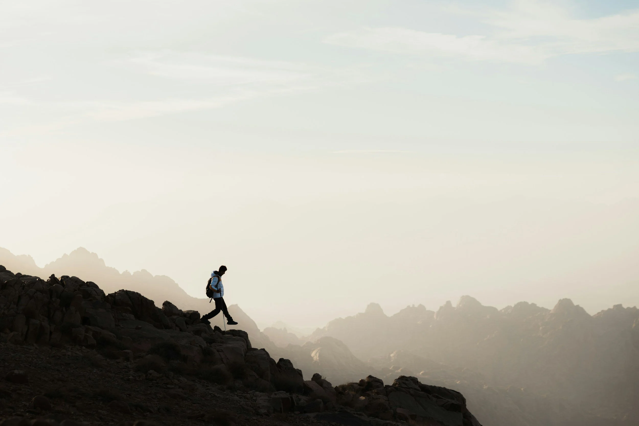 A person hiking on a rocky mountain trail during daytime, with mountains and a cloudy sky in the background.
