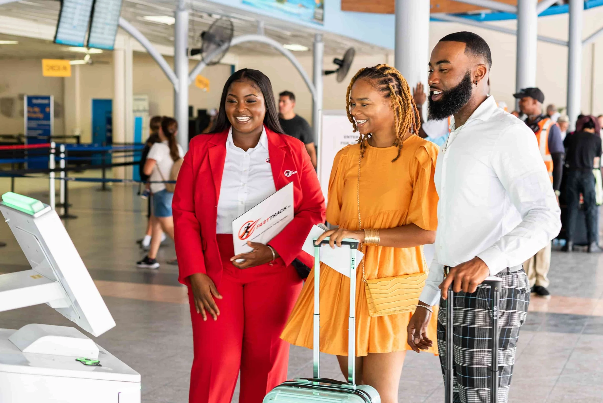 A woman in a red suit and two people with a yellow dress and plaid pants are at an airport check-in kiosk, smiling and talking.