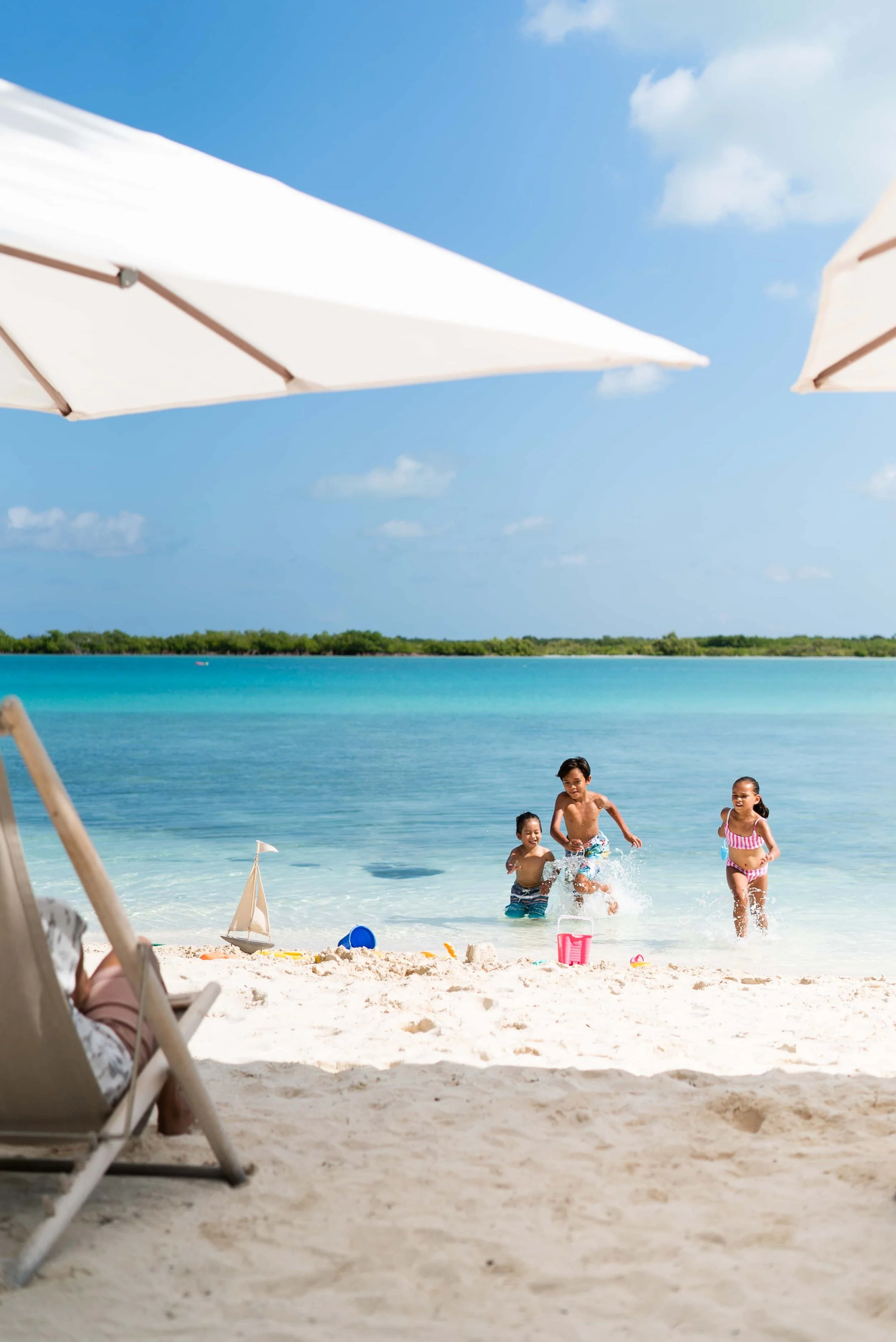 Children playing and splashing in the ocean water on a sunny day at the beach, with umbrellas and sand toys nearby.