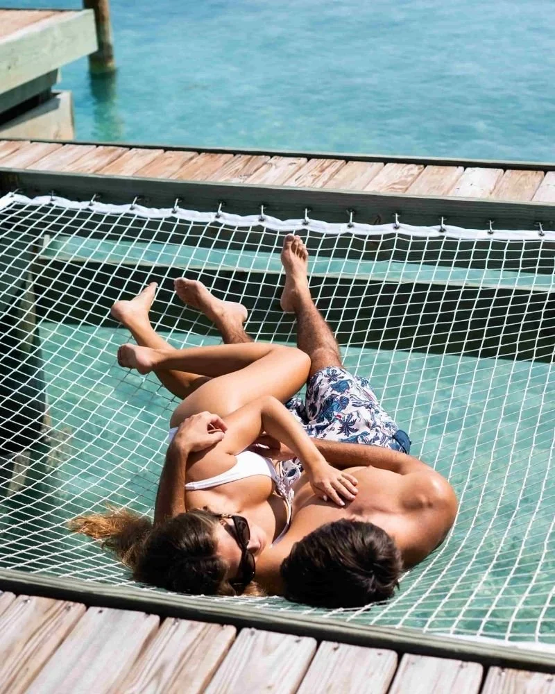 Two people lying in a hammock over water on a dock, with one person wearing sunglasses and a swimsuit, and the other in shorts.