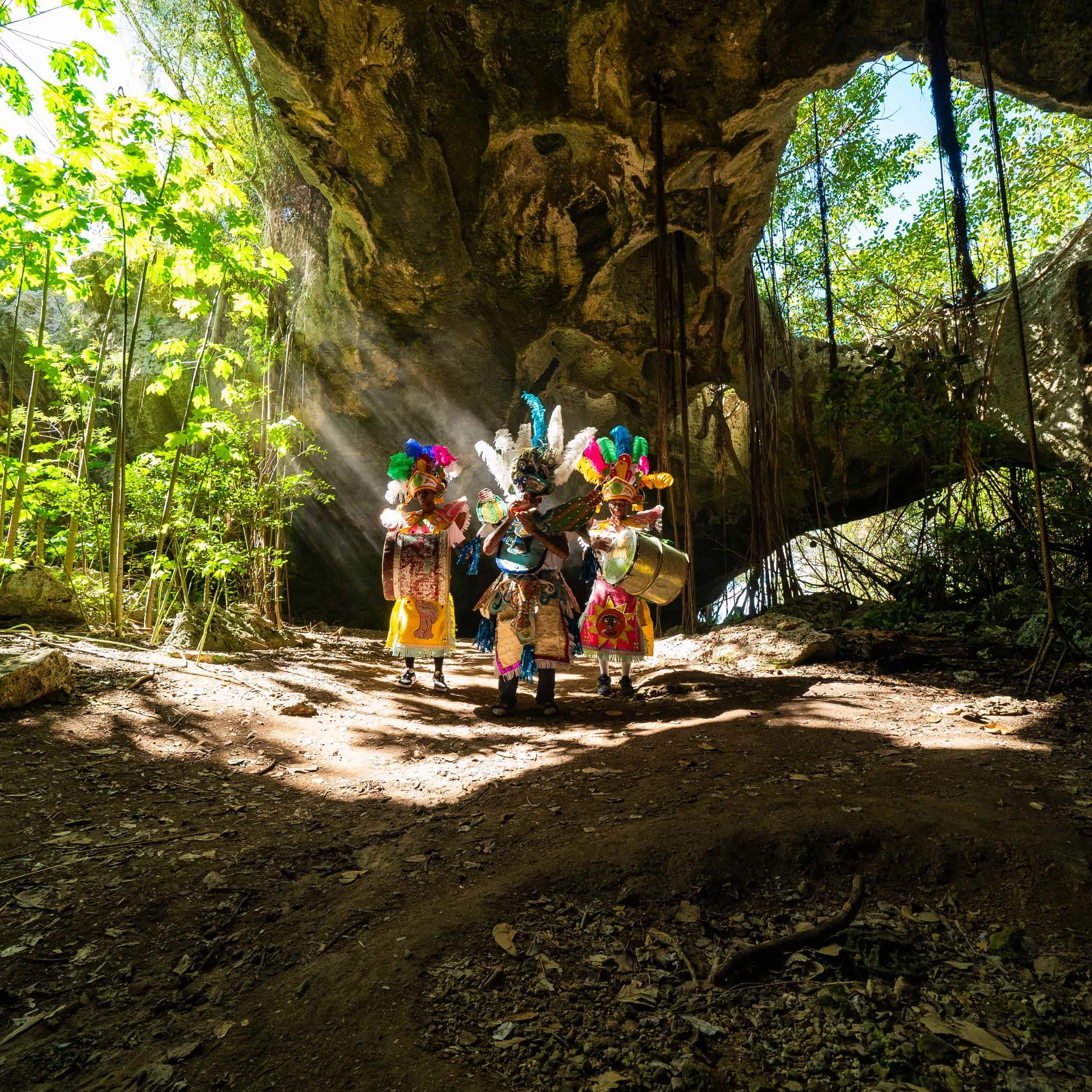 Indigenous dancers in colorful traditional costumes and feathered headdresses perform inside a rocky cave with sunlight streaming through openings, surrounded by lush green foliage.