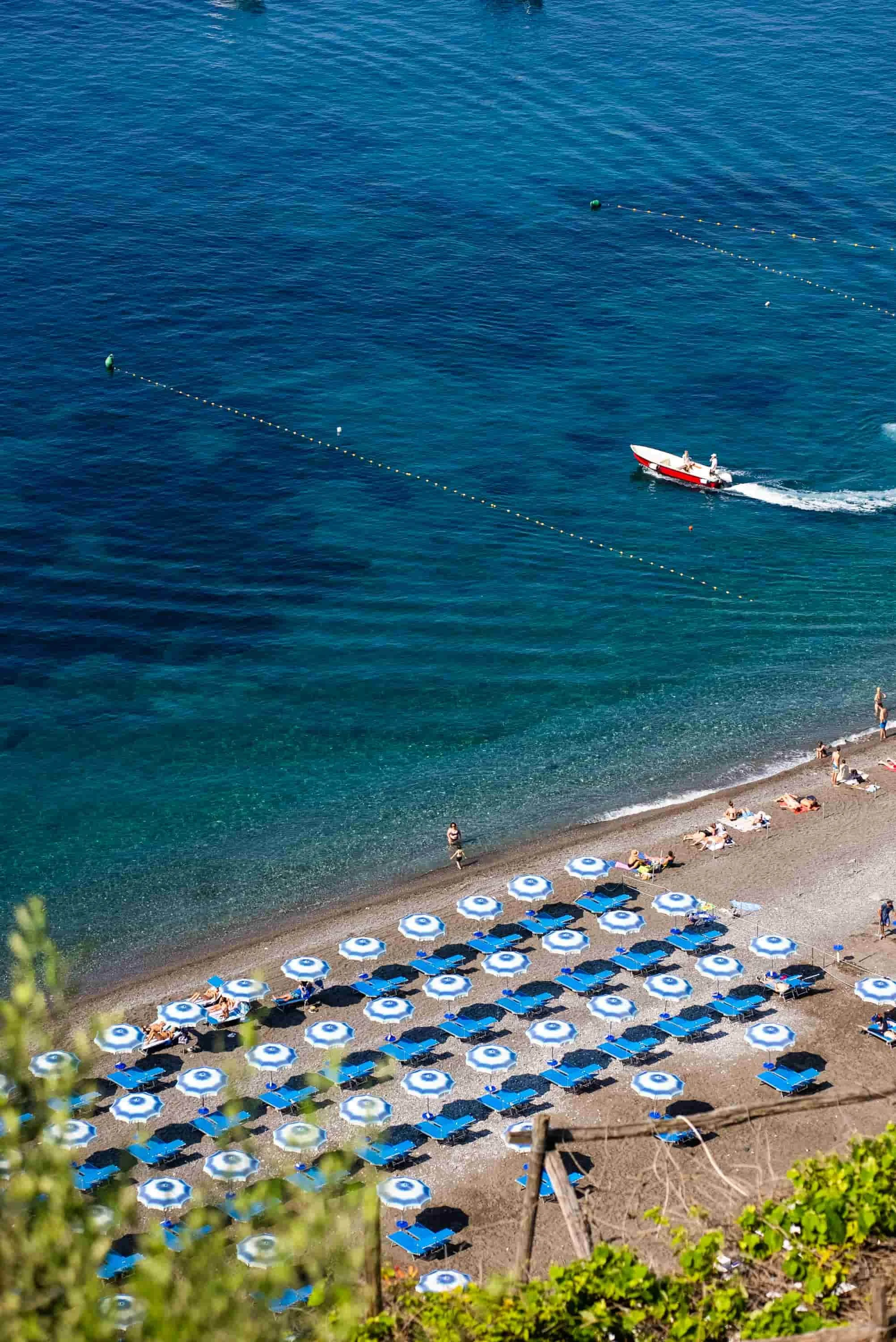 Beach with blue umbrellas and lounge chairs, people sunbathing and walking along the shore, a boat with passengers, and a calm, blue sea with buoys and a moving boat.
