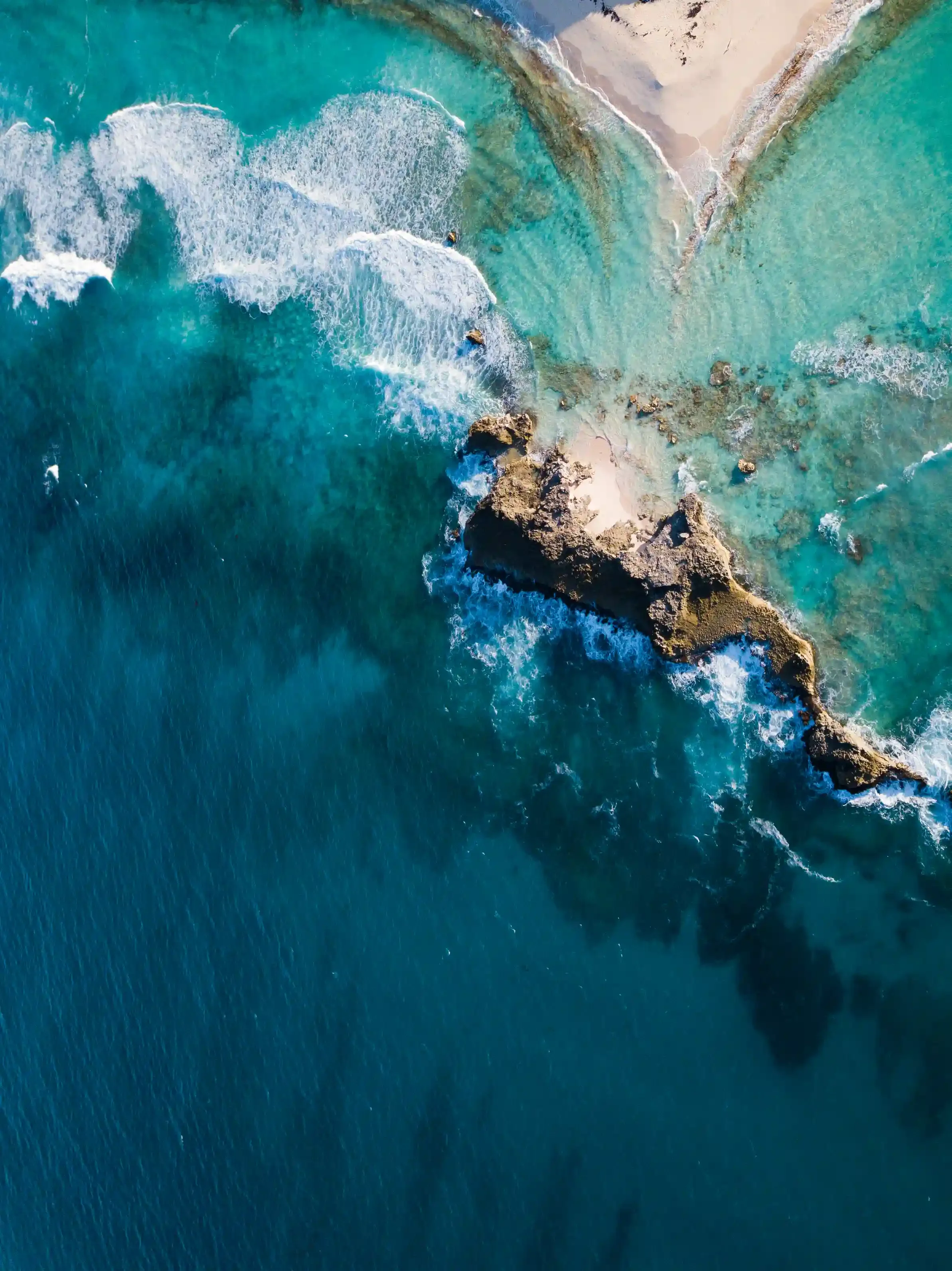 Aerial view of a rocky coast with turquoise waters, waves crashing against rocks and sandy shoreline.