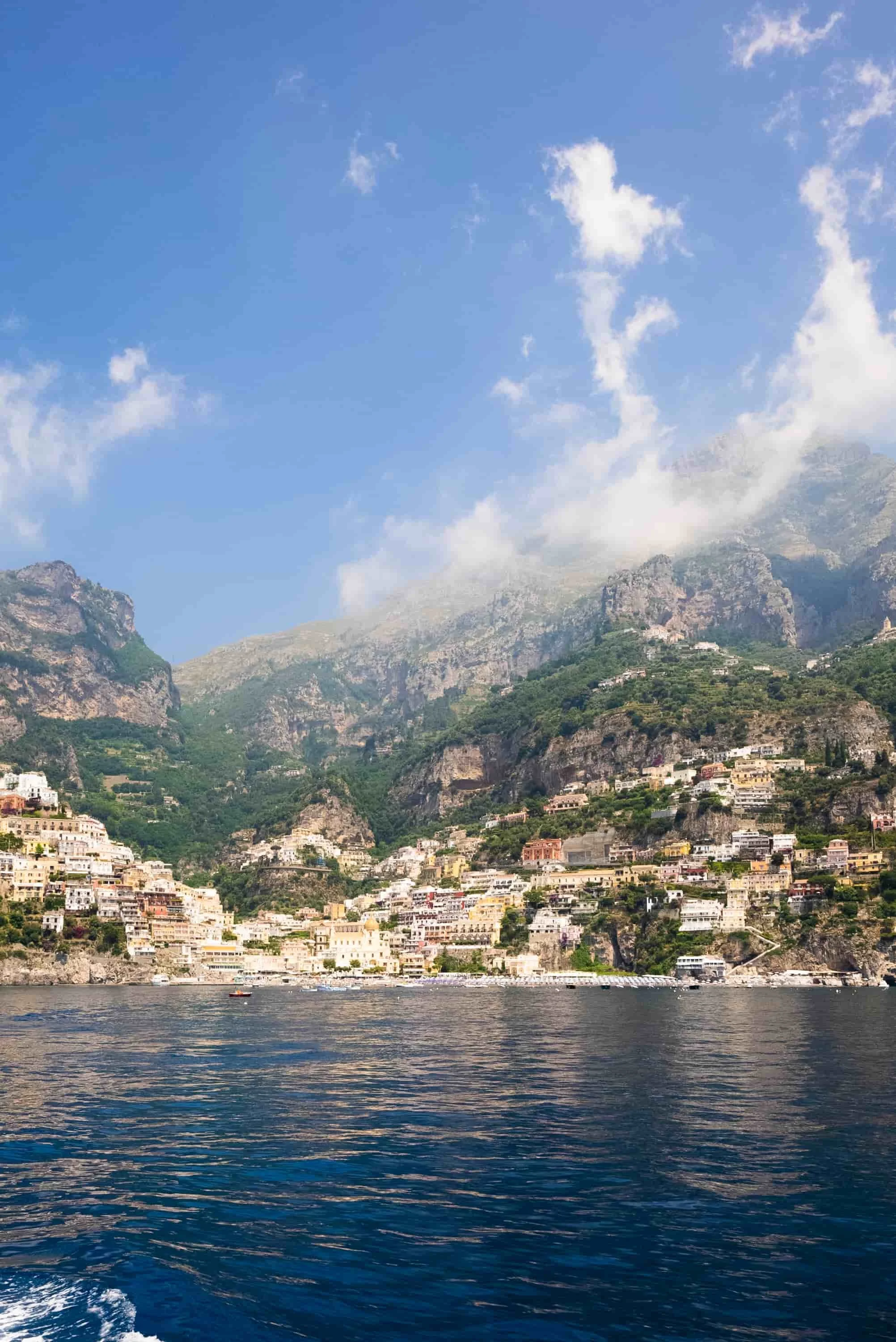 Scenic view of a coastal town built on steep hillsides with colorful buildings, overlooking calm dark blue water, with mountains and partly cloudy sky in the background.