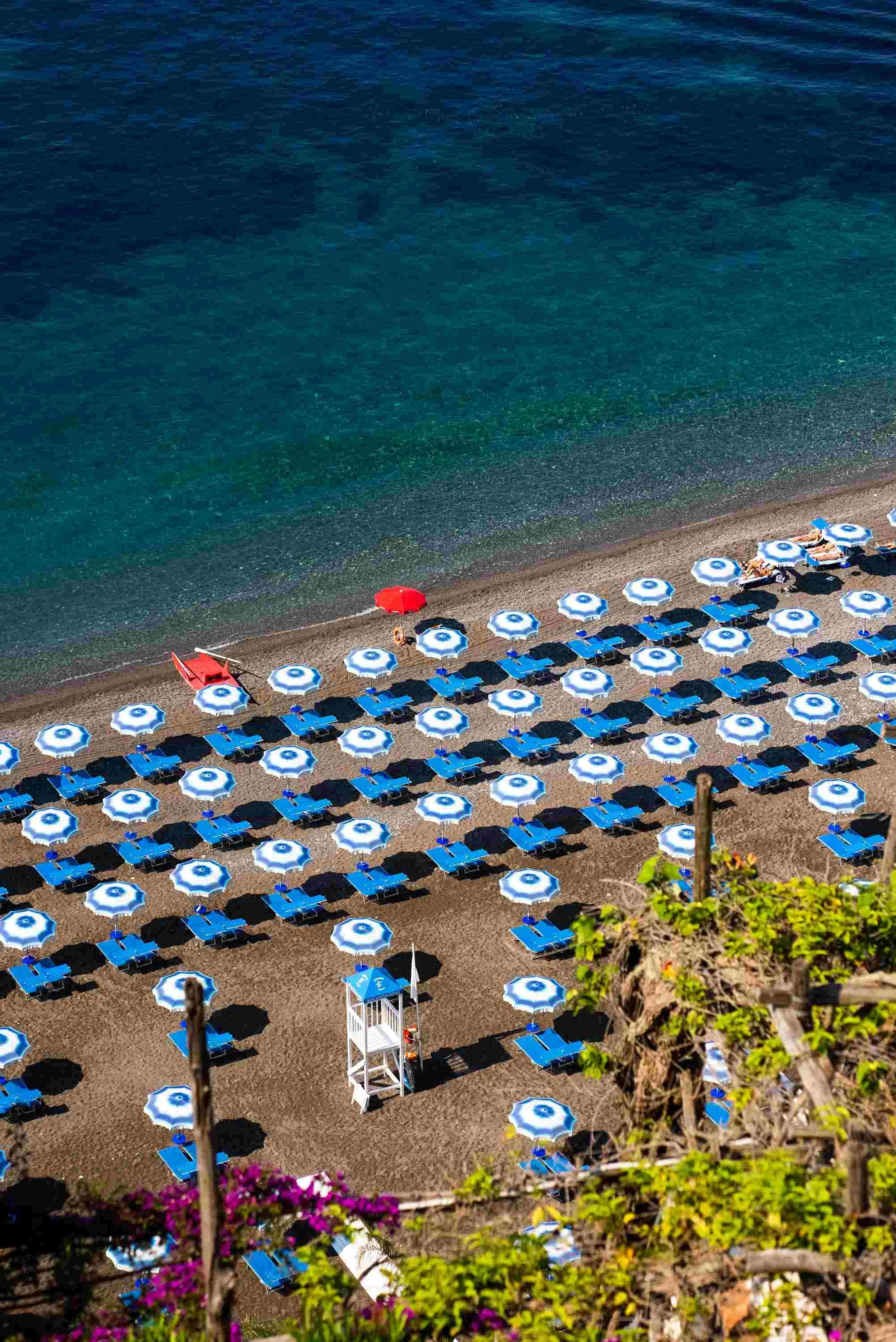 Empty beach with blue and white striped umbrellas, blue sun loungers, a white lifeguard tower, and a few people lounging under umbrellas, overlooking calm blue sea.