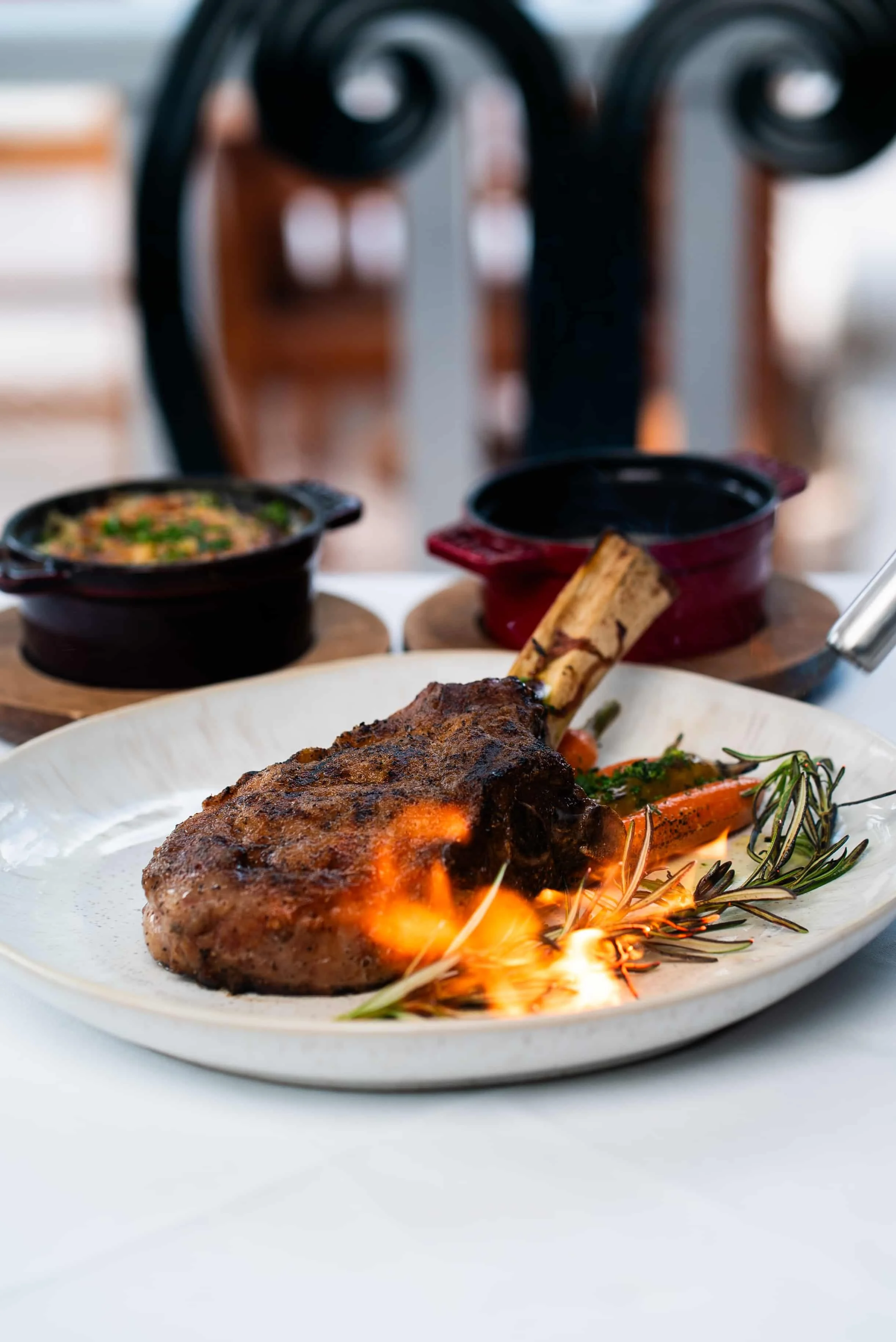 A roasted meat dish with a bone, garnished with herbs and vegetables, presented on a white plate, with flames underneath.