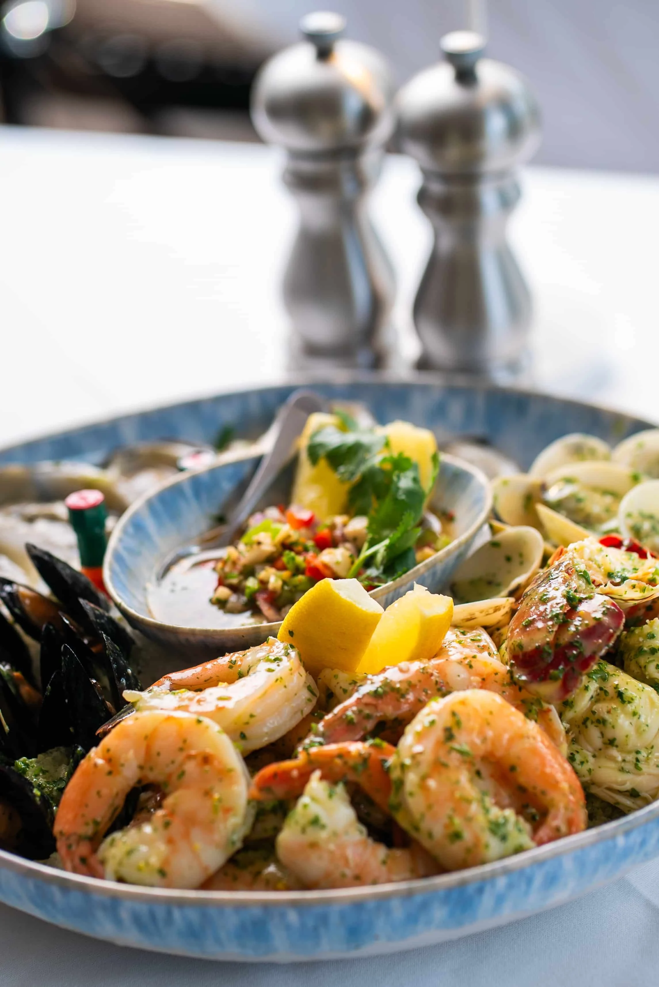 Seafood platter with shrimp, mussels, and clam linguine, garnished with lemon wedges and fresh herbs on a blue ceramic plate, with salt and pepper shakers in the background.