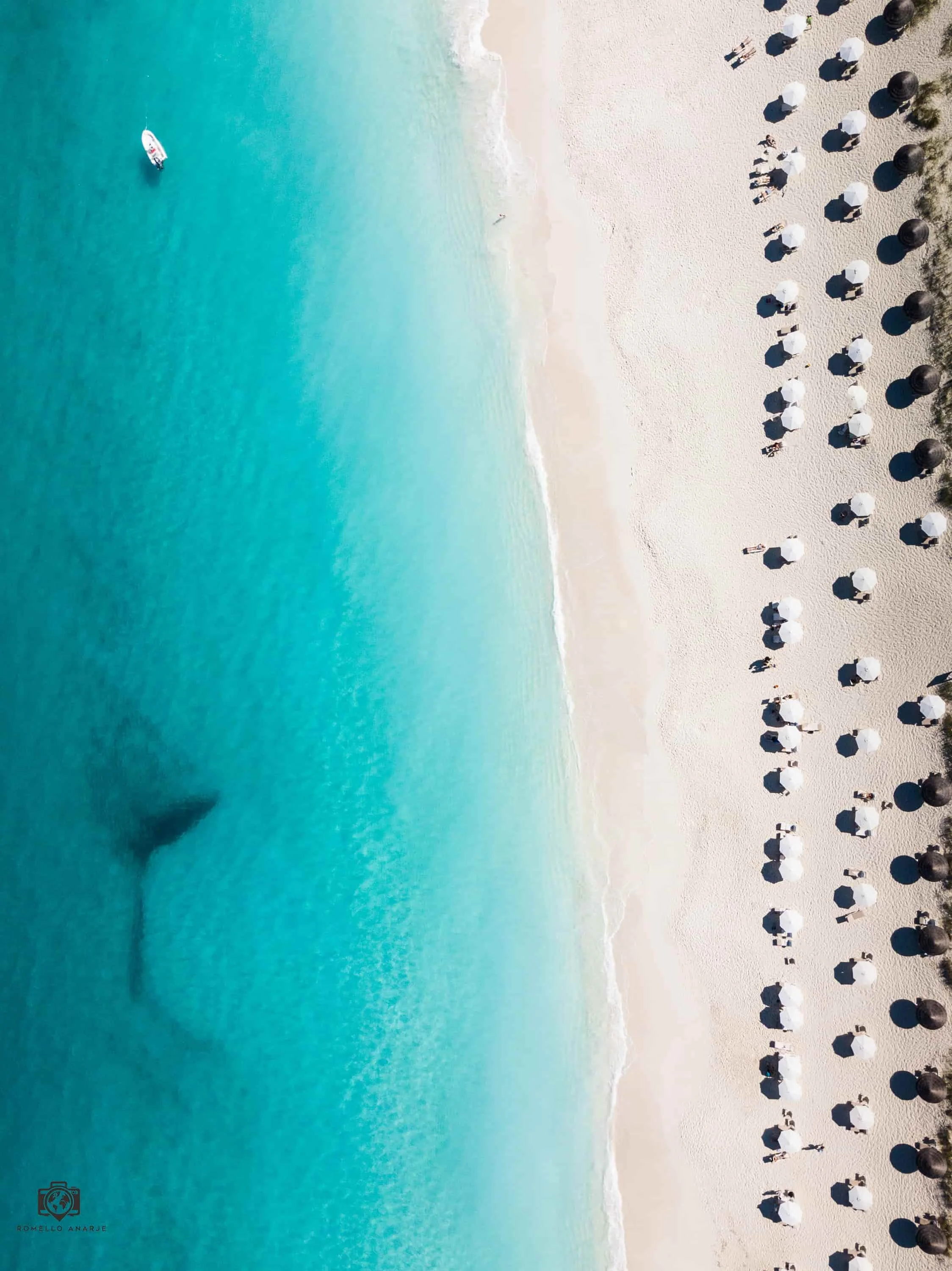 An aerial view of a beach with clear turquoise water and a boat, lined with umbrellas and people walking on the sand.