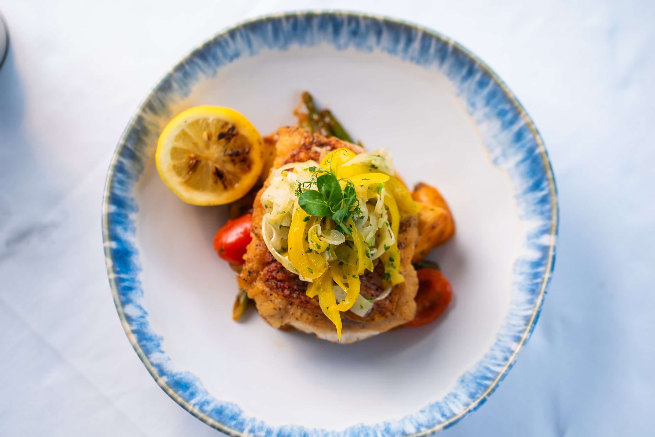 A plate with fried fish topped with sautéed vegetables and garnished with fresh herbs, accompanied by a halved lemon on a decorative blue and white rimmed plate.
